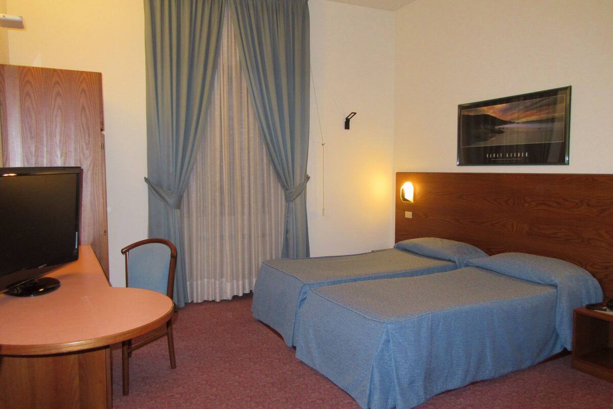 A hotel room with two beds, a desk with a television, a chair, and a wall-mounted light.