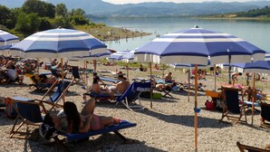 A beach with multiple blue and white striped umbrellas, people relaxing on sun loungers, and a view of a lake and mountains in the background. - Hotel Giardino (Prato)