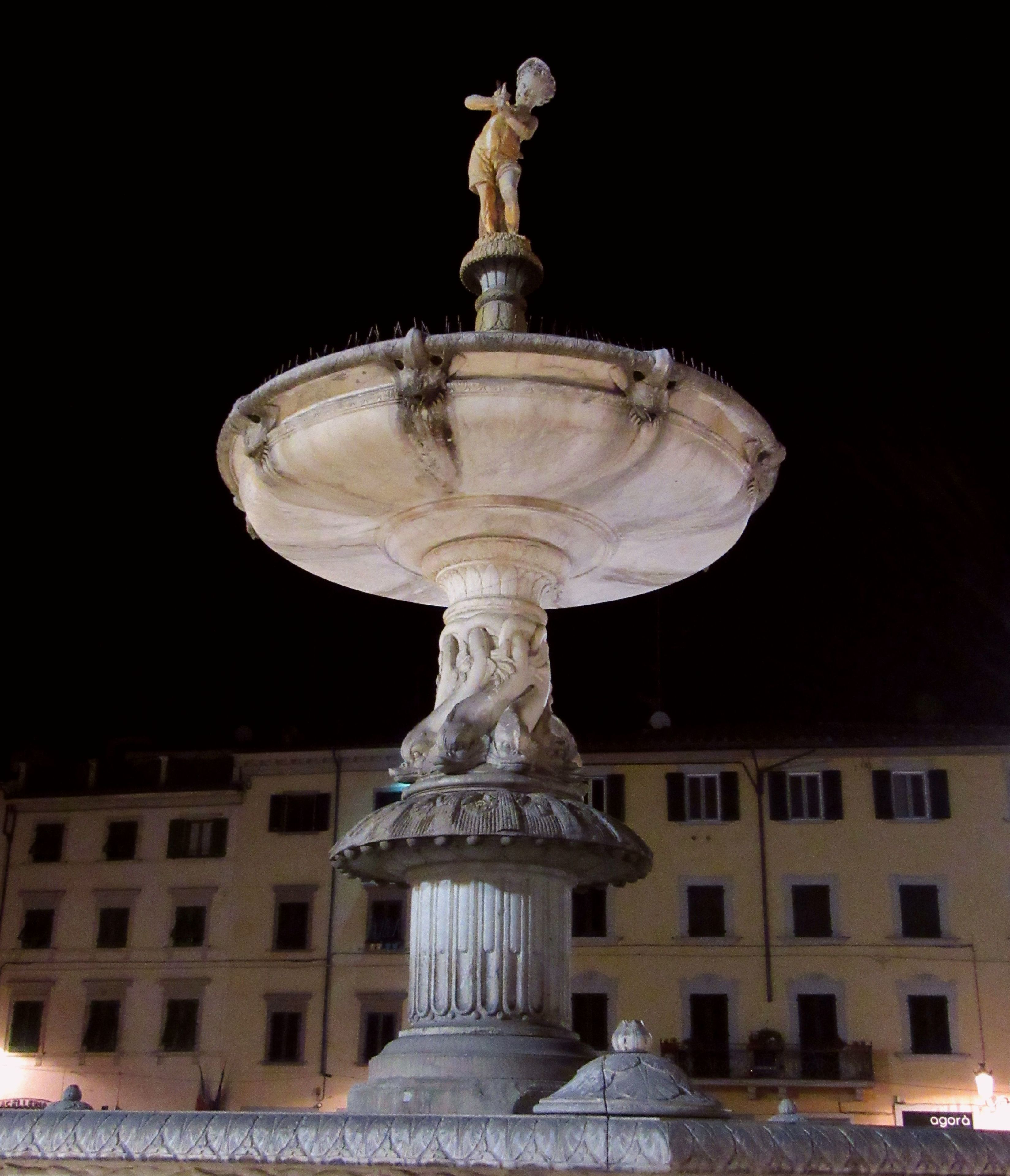 A night-time fountain with a statue on top, set against a building with multiple windows.