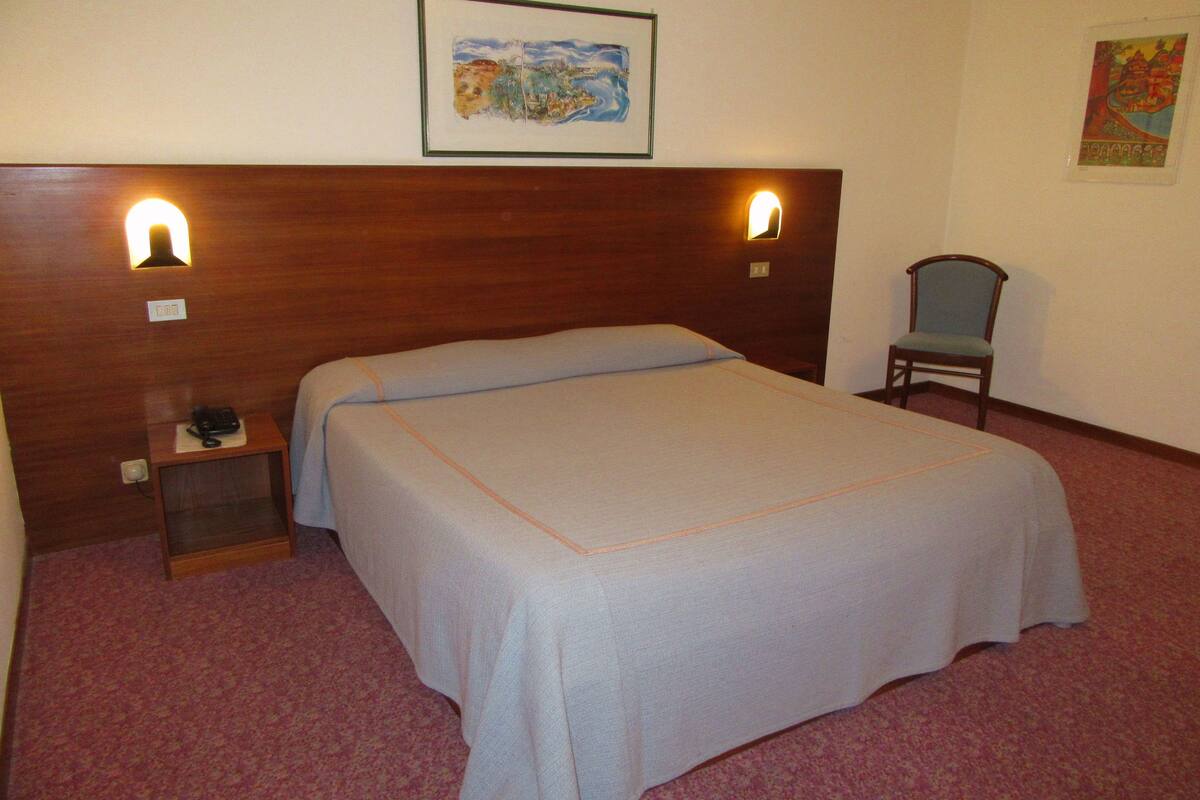 A hotel room with a bed, a nightstand with a phone, a chair, and two wall-mounted lights.