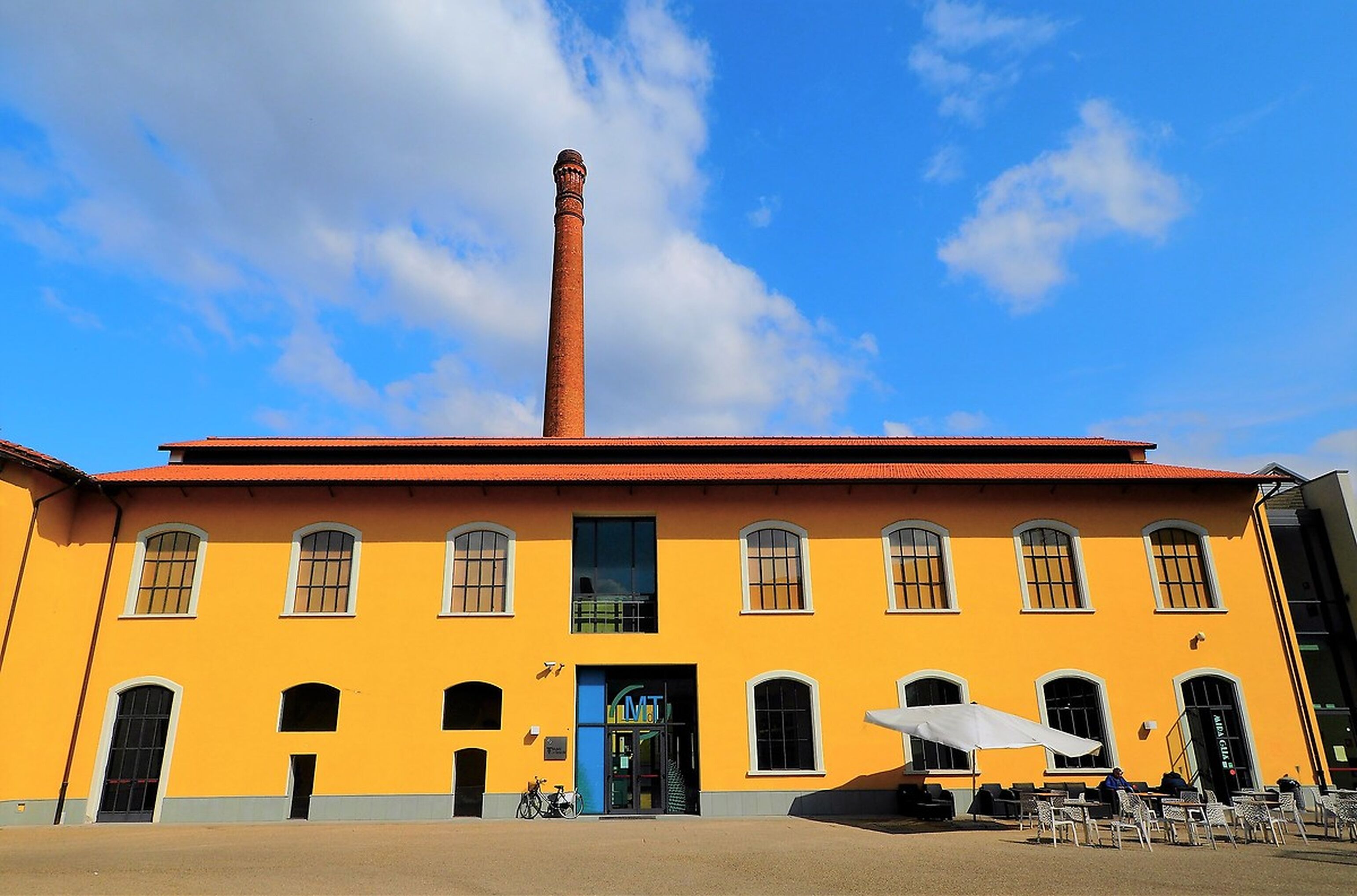 Ein gelbes Gebäude mit einem hohen Schornstein, Bogenfenstern und Außenbestuhlung mit Sonnenschirmen.