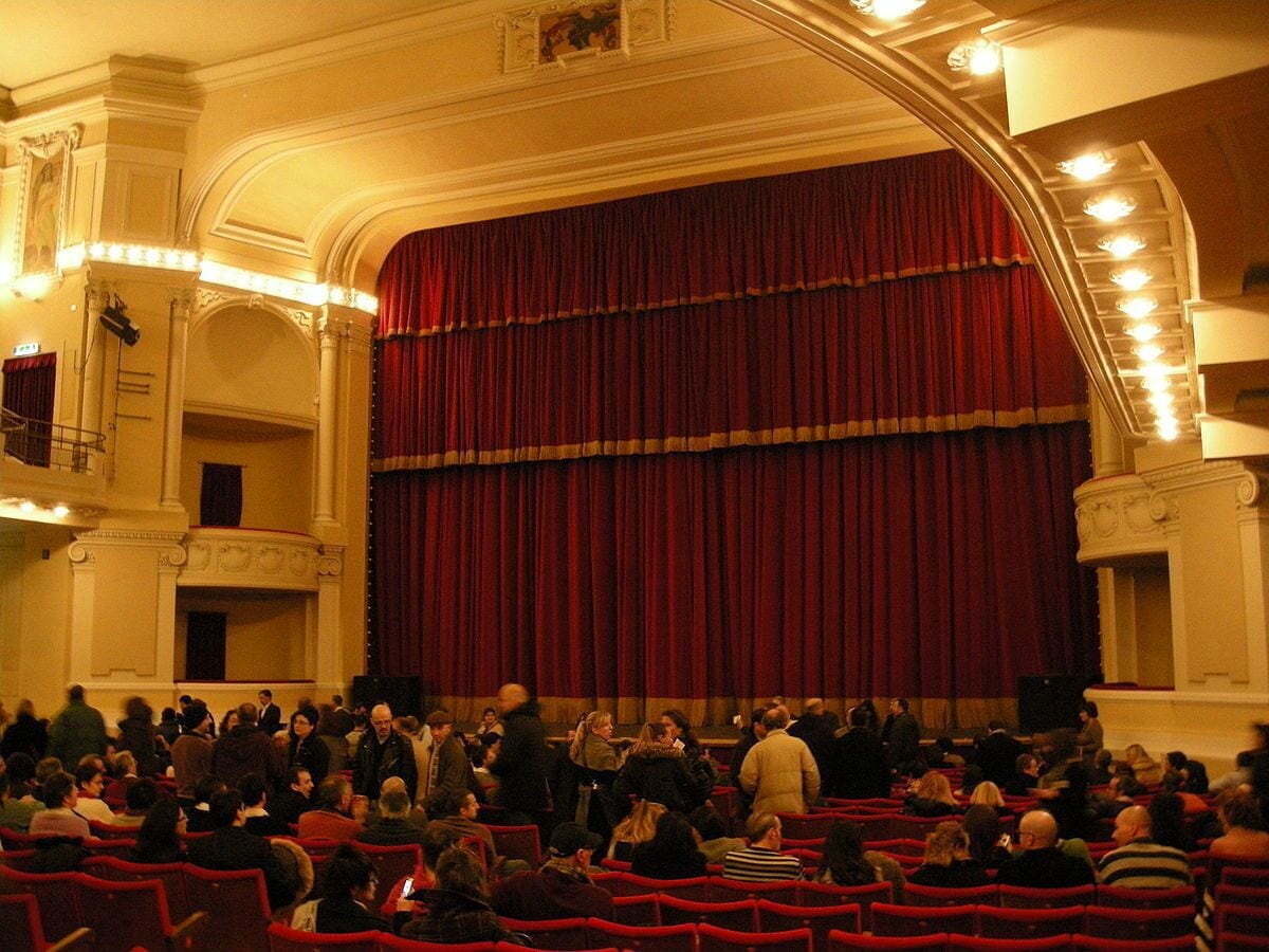 A theater with a large audience seated in red chairs, a stage with a red curtain, and ornate architectural details.