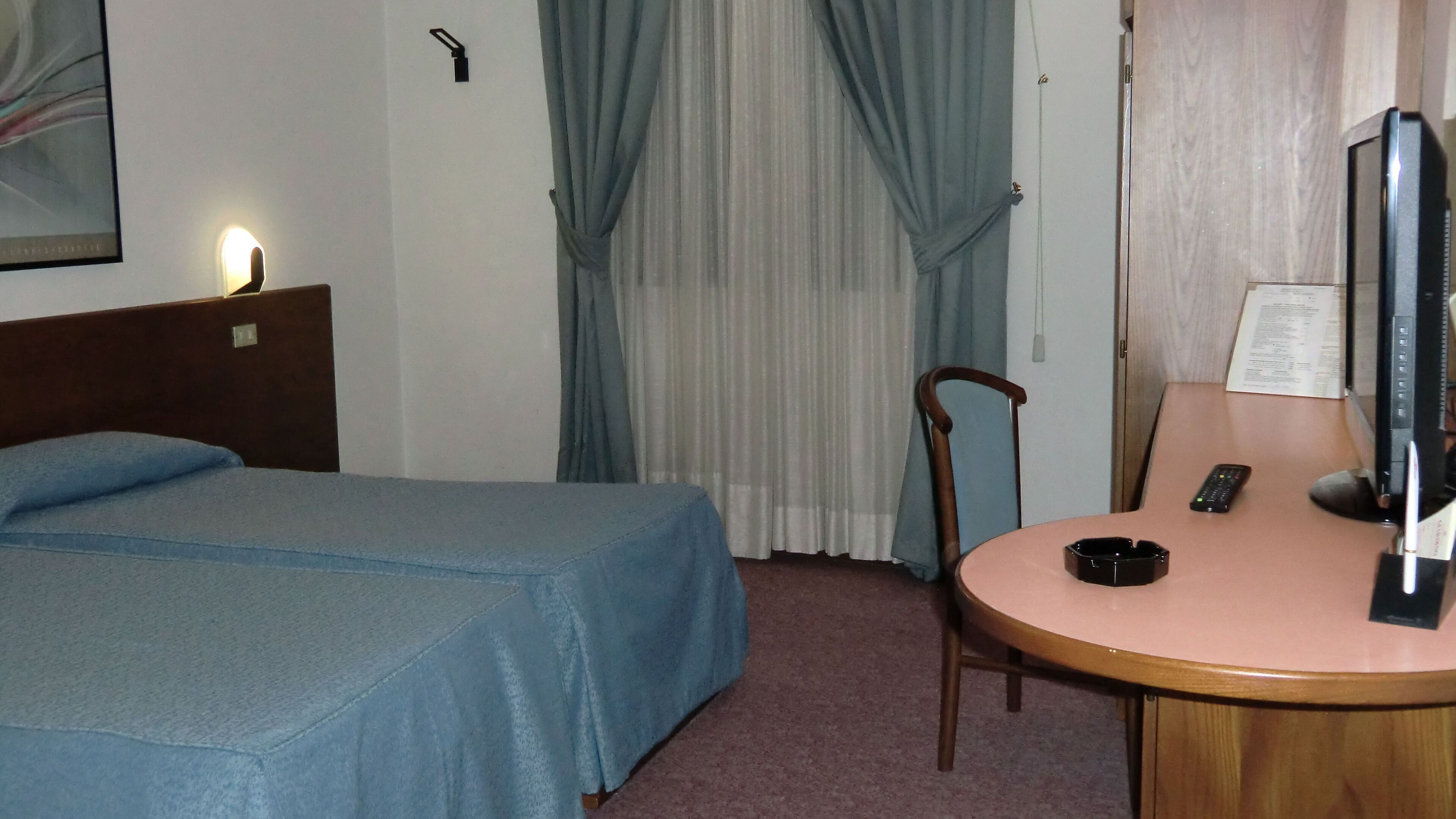 A hotel room with a bed, a desk with a computer, a chair, a television, and a window with curtains.