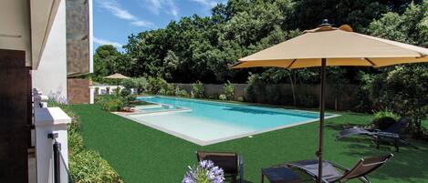 Una piscina con zona de patio, que incluye una mesa y sillas, rodeada de follaje y una valla.