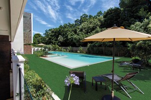 Una piscina con zona de patio, que incluye una mesa y sillas, rodeada de follaje y una valla.