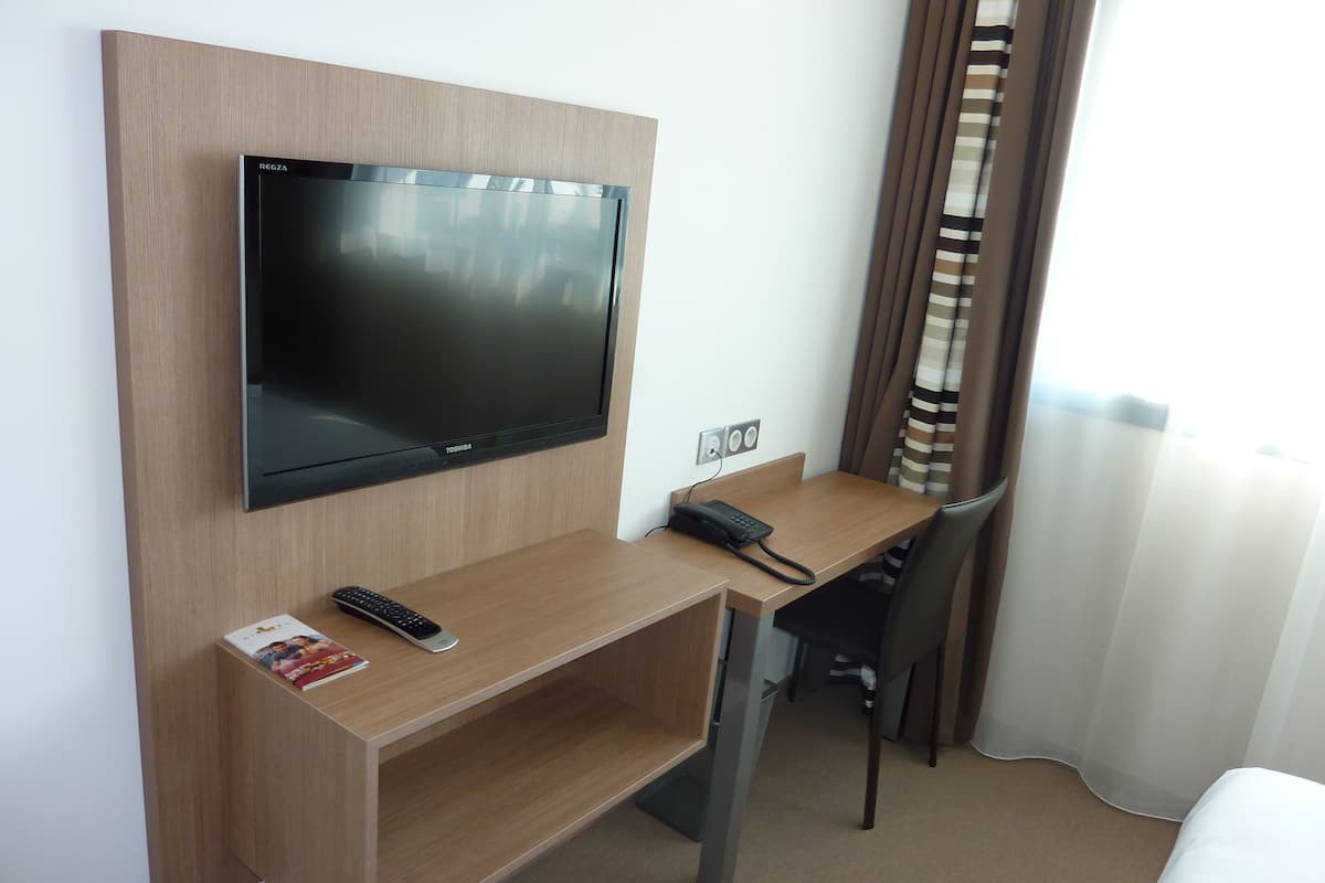Ein Hotelzimmer mit einem an der Wand befestigten Fernseher, einem Schreibtisch mit Telefon und einem Sessel.