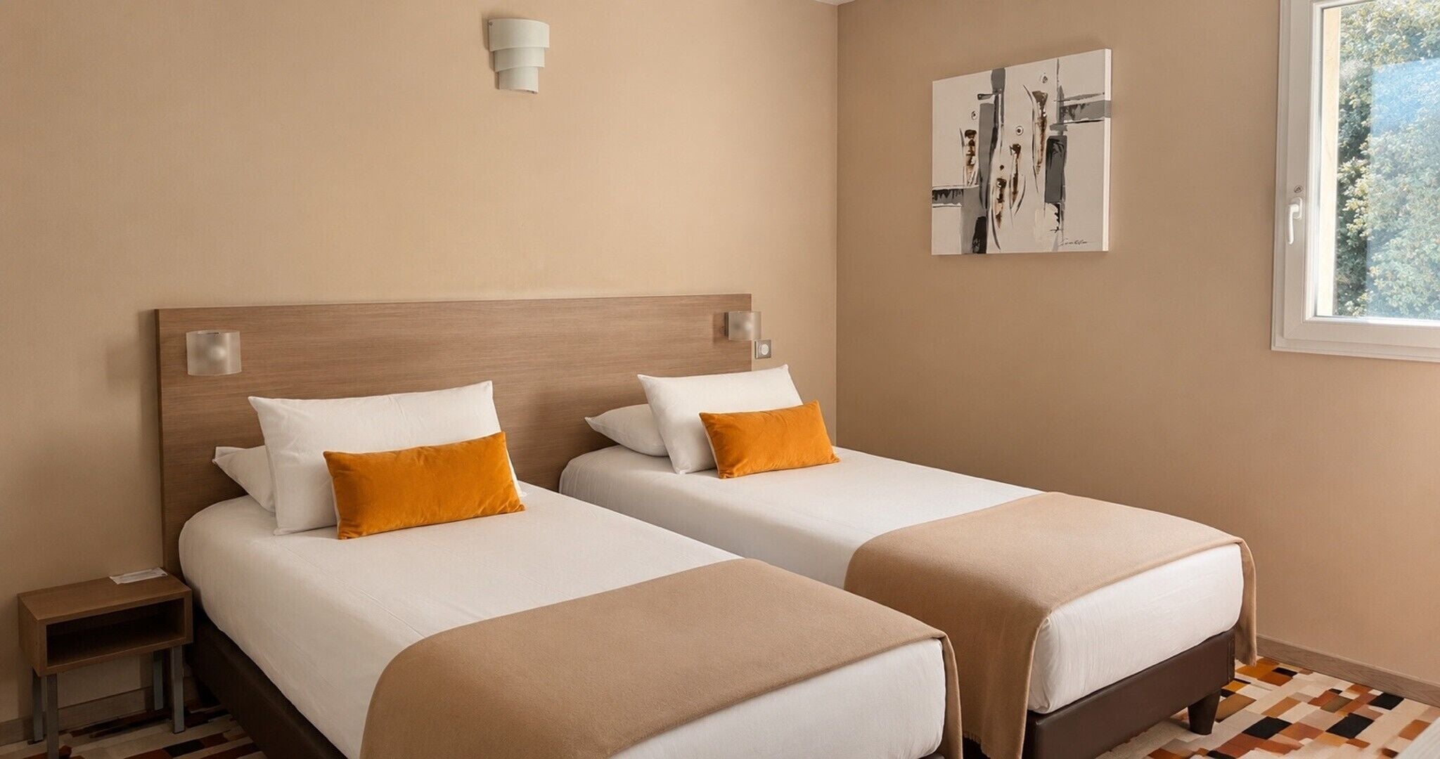 A hotel room with two beds, a nightstand, a wall-mounted light, and a window with a view.