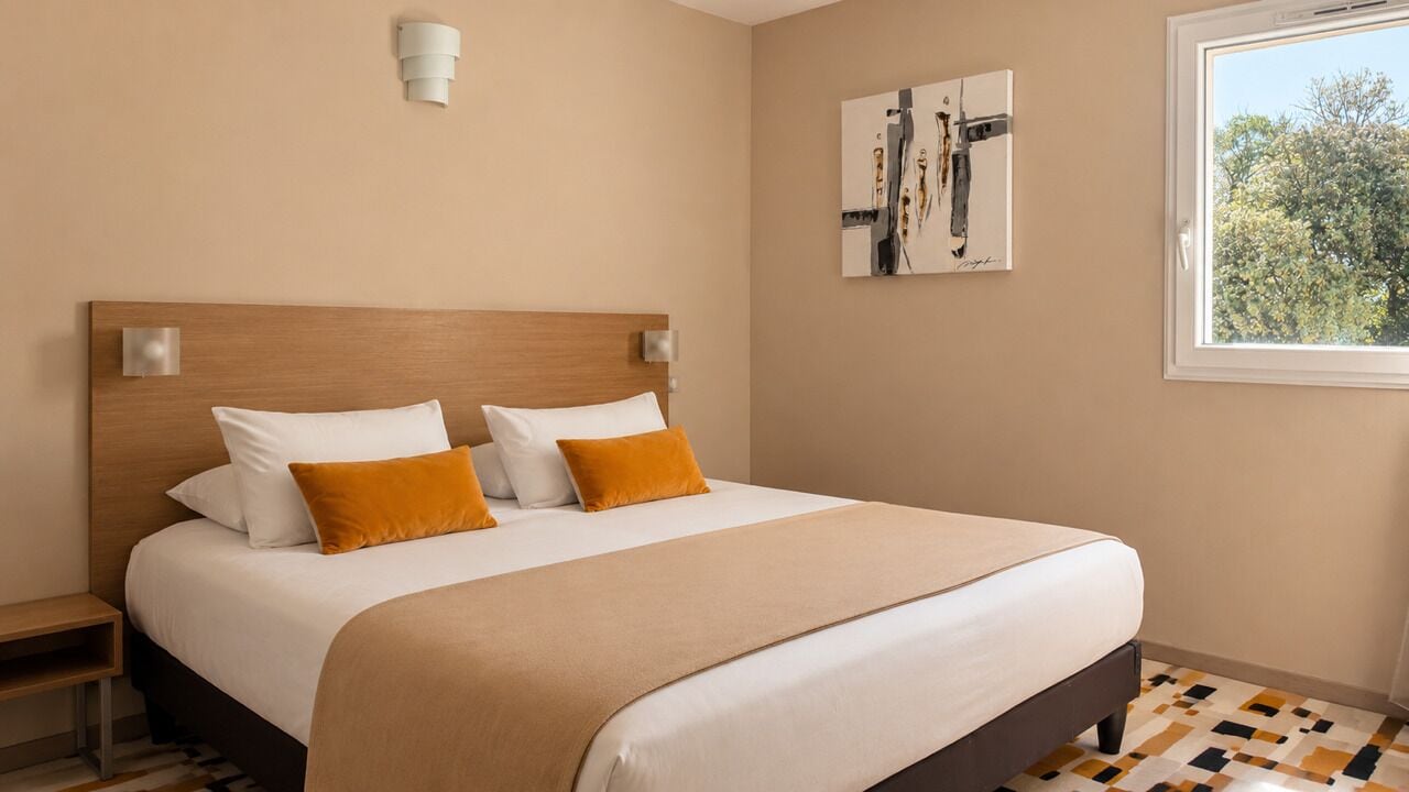 Un dormitorio con cama, mesita de noche, lámpara de pared y ventana con vista a árboles.