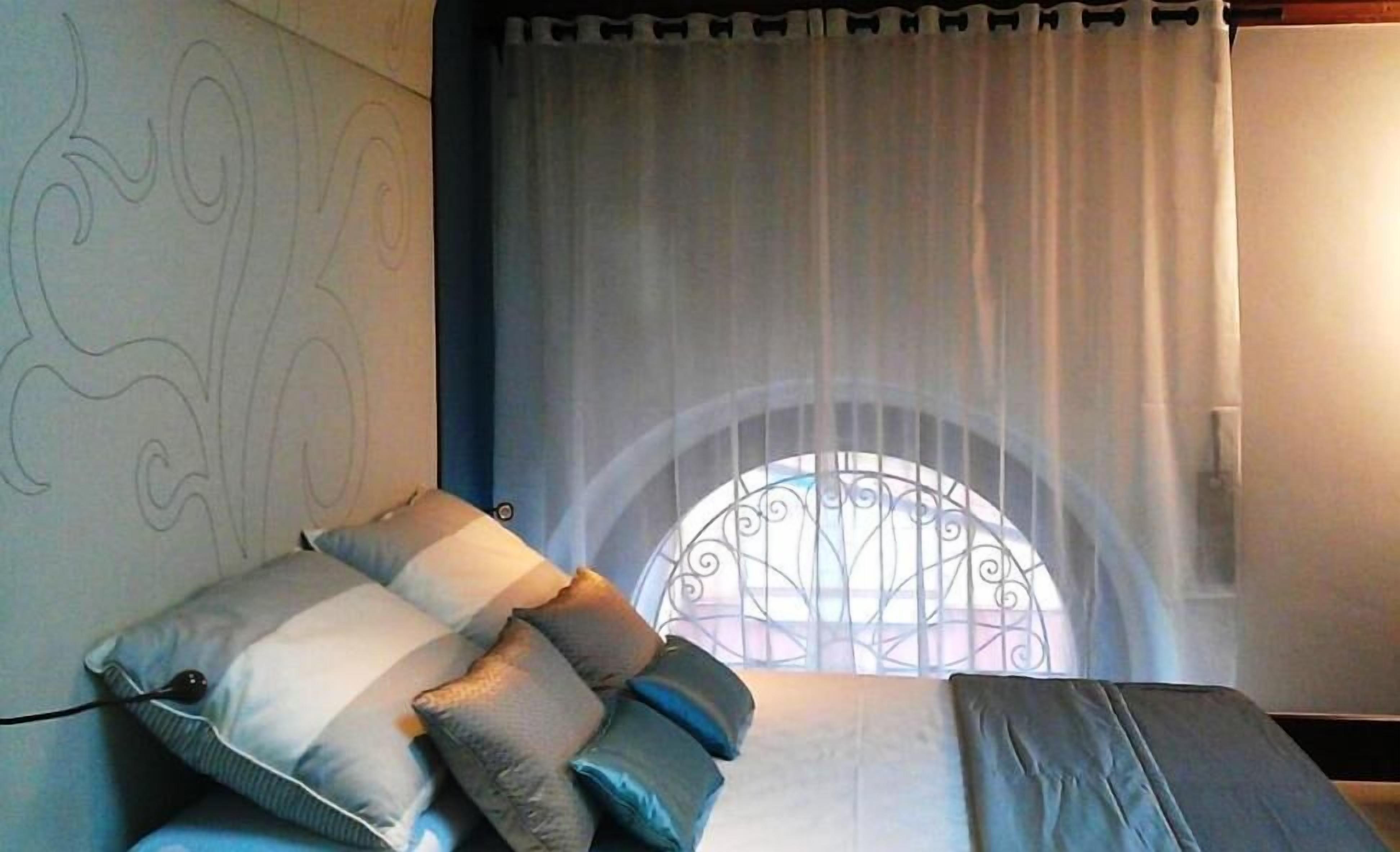 Un dormitorio con cama, almohadas, una cortina y una ventana con reja de metal.