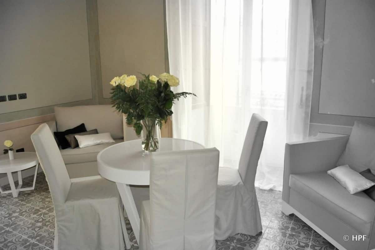Een moderne, minimalistische woonkamer met een ronde witte tafel, twee witte stoelen, een witte bank en een vaas met gele bloemen.