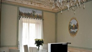 Een hotelkamer met een televisie op een standaard, een bank, een eettafel met stoelen, een kroonluchter en een schilderij aan de muur.