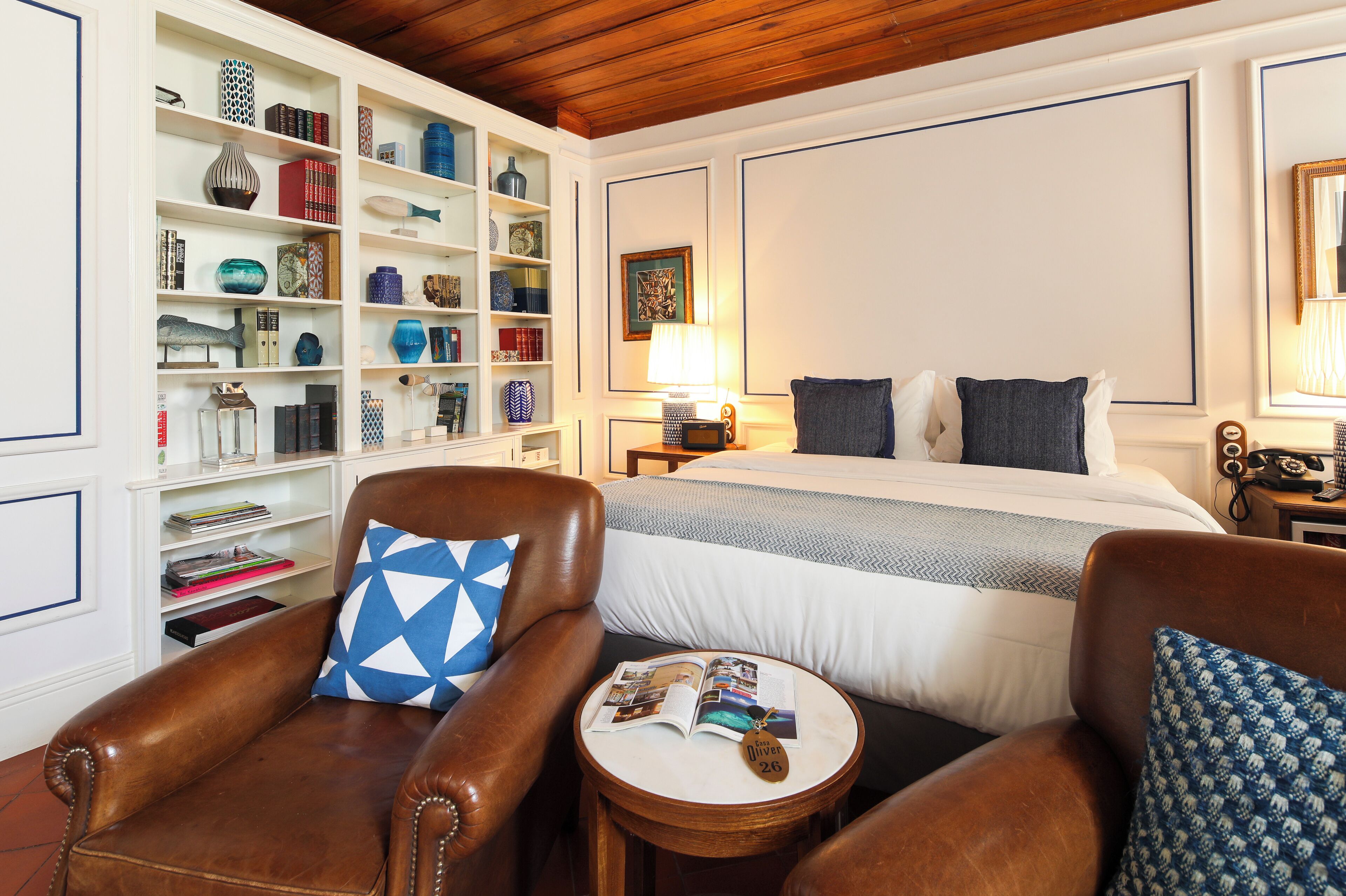 Een hotelkamer met een groot bed, een leren fauteuil, een klein rond tafeltje met een tijdschrift, en een boekenplank vol boeken en decoratieve objecten.