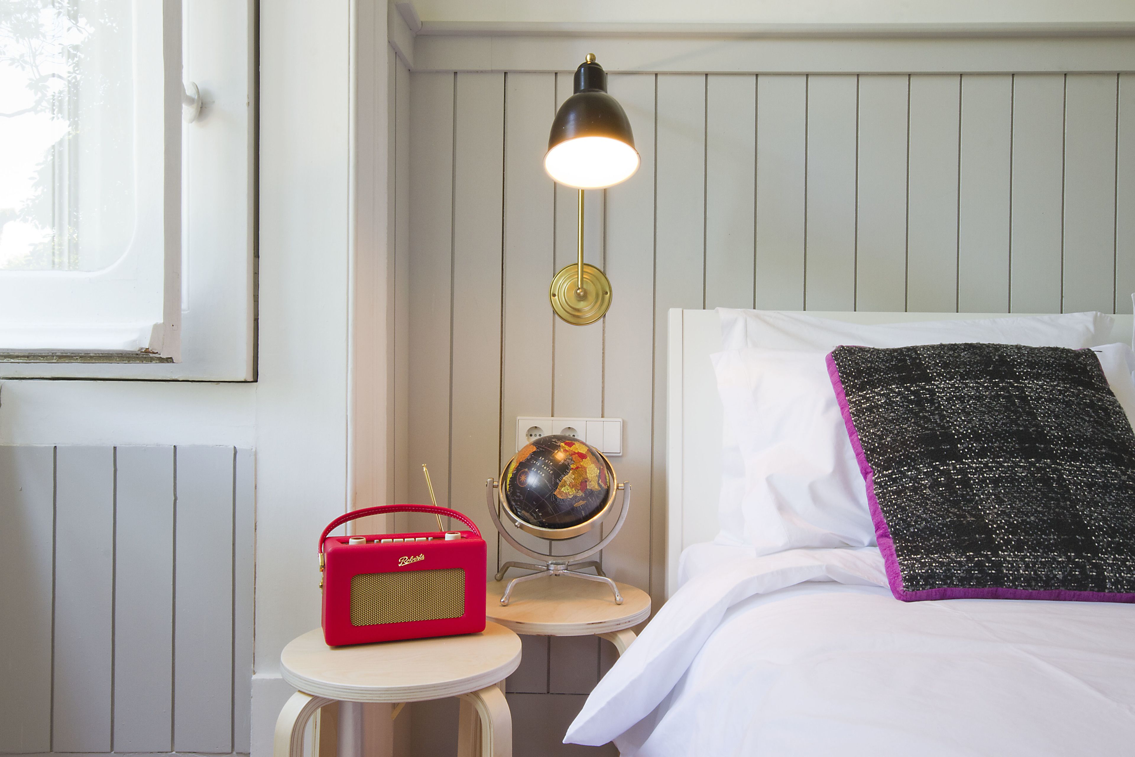 Un dormitorio ordenado con una cama, un globo terráqueo sobre un atril, una radio roja y una lámpara en una mesita de noche.
