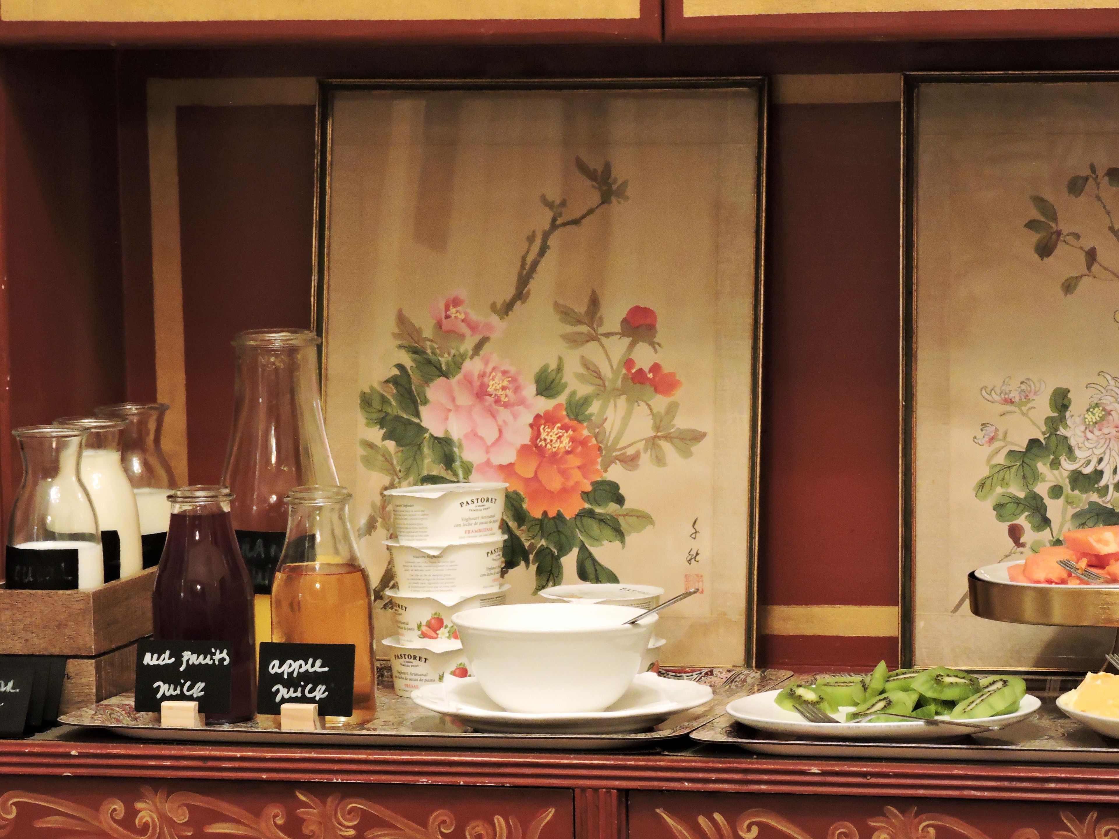 A breakfast buffet with various food items and beverages, including red fruits juice and apple juice, and a traditional painting in the background.