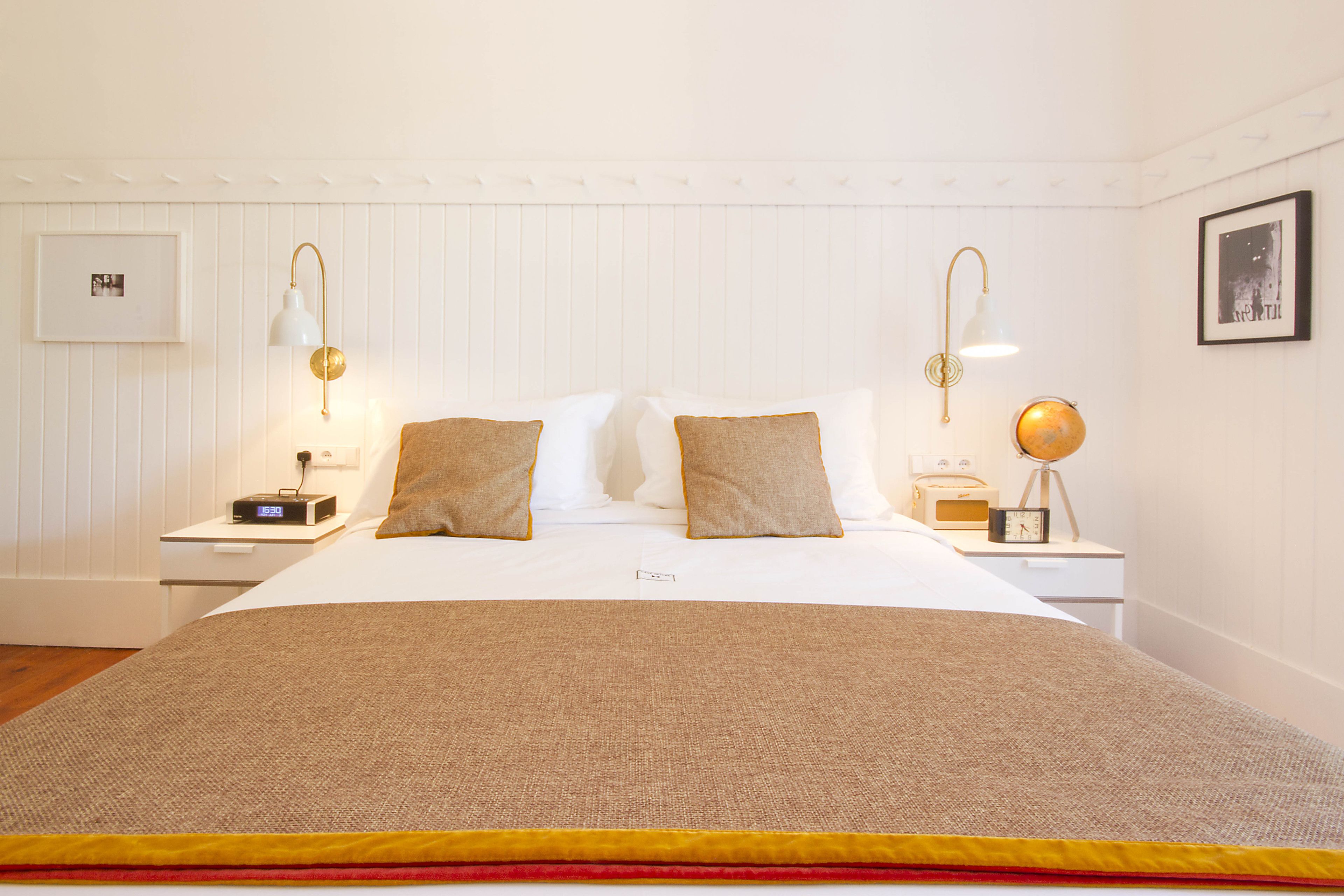 Een net opgemaakt bed met een bruine deken en kussens, twee nachtkastjes met lampen, een ingelijste foto aan de muur, en een wereldbol op een van de tafels.