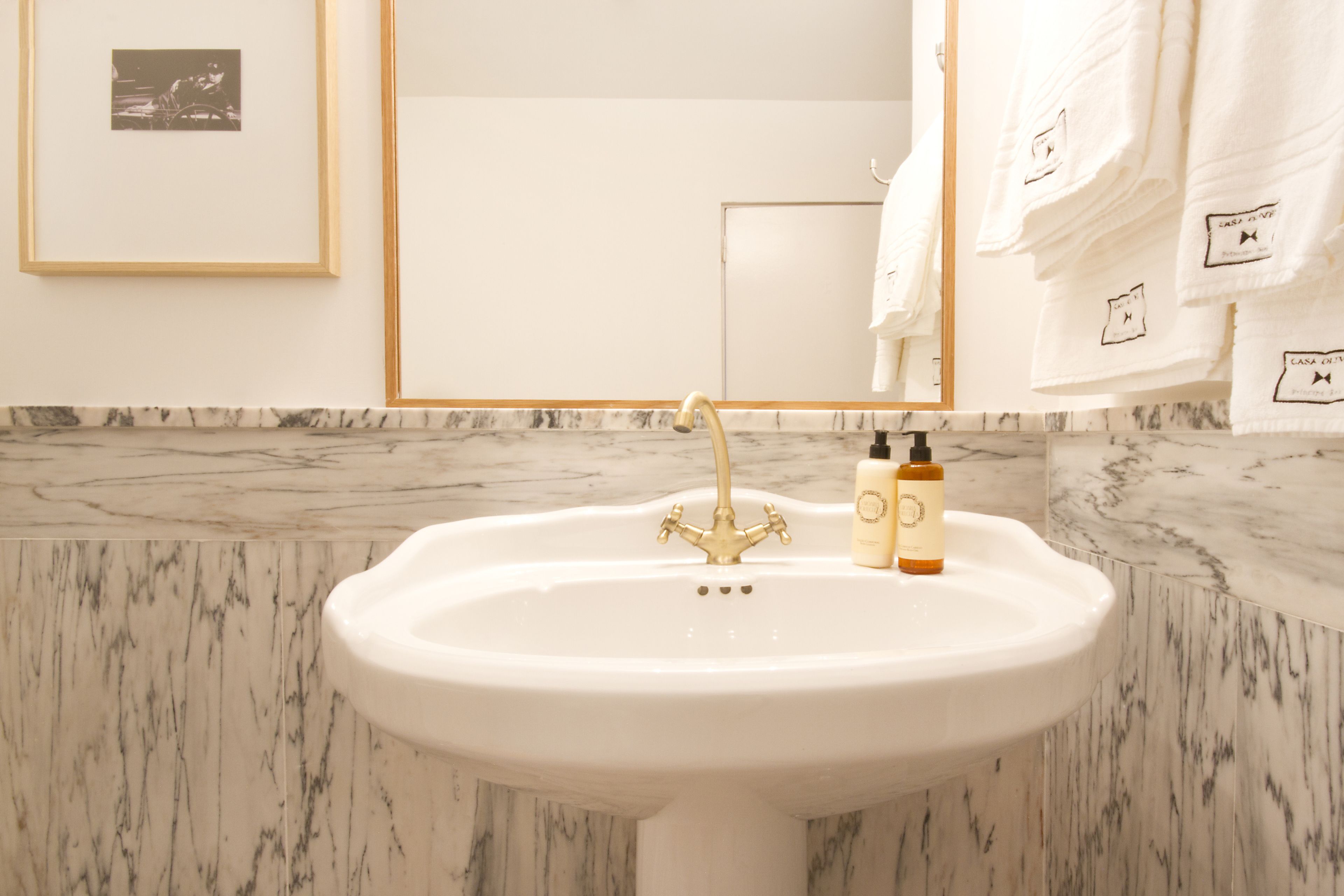 A bathroom with a white sink, gold faucet, and marble backsplash. There are two bottles on the sink and towels hanging on the wall.