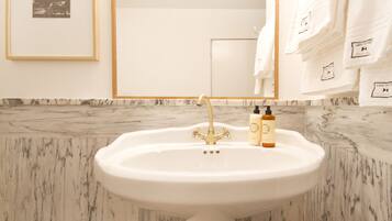 A bathroom with a white sink, gold faucet, and marble backsplash. There are two bottles on the sink and towels hanging on the wall.