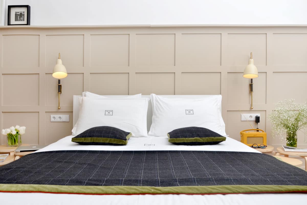 A neatly made bed with a dark checkered bedspread, two white pillows, and two dark pillows with green trim. There are two bedside lamps with beige shades, a small table with a vase of flowers, and a yellow radio.
