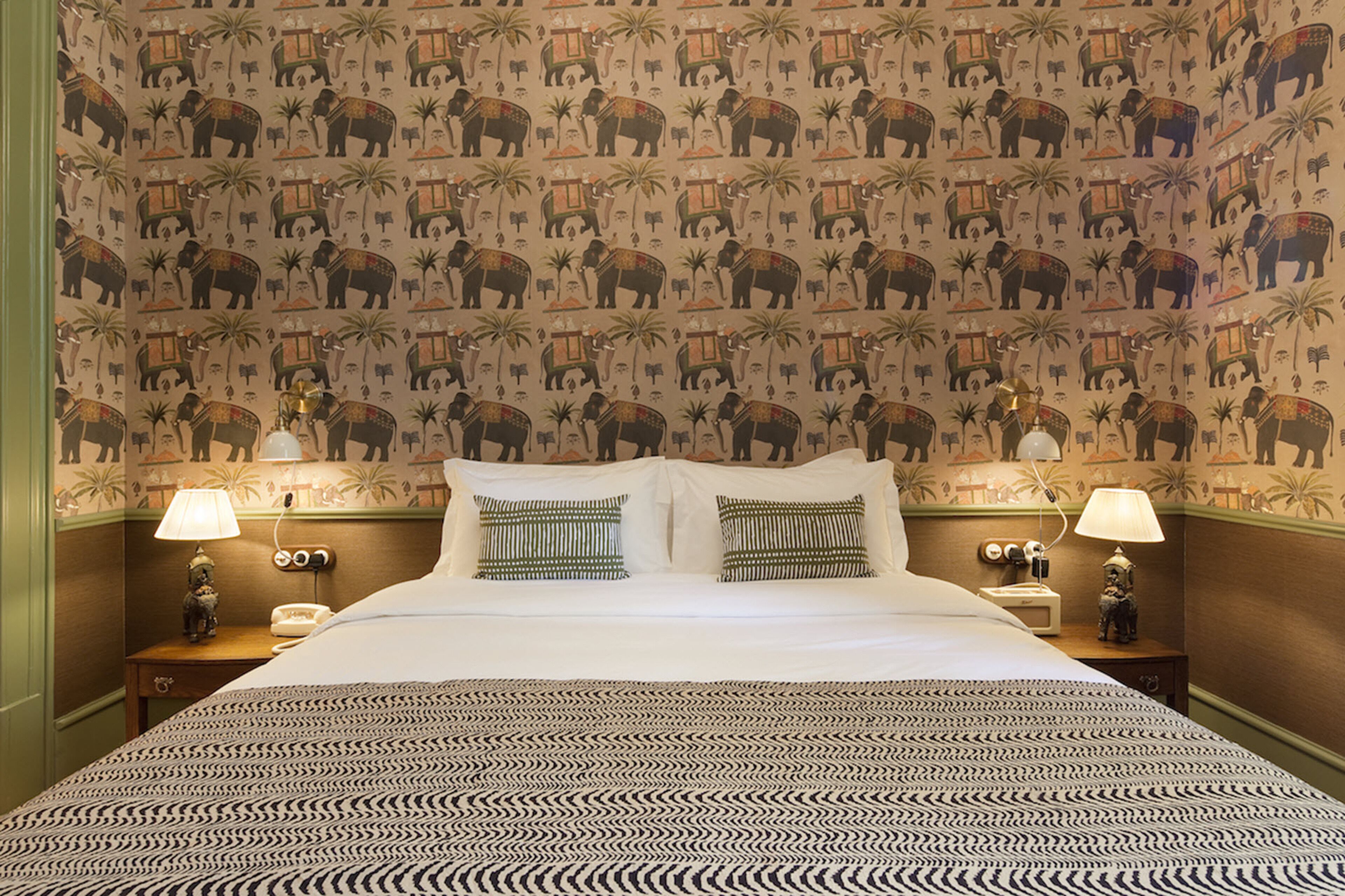 A hotel room with a patterned wallpaper, a bed with white linens, two bedside lamps, and a patterned bedspread.