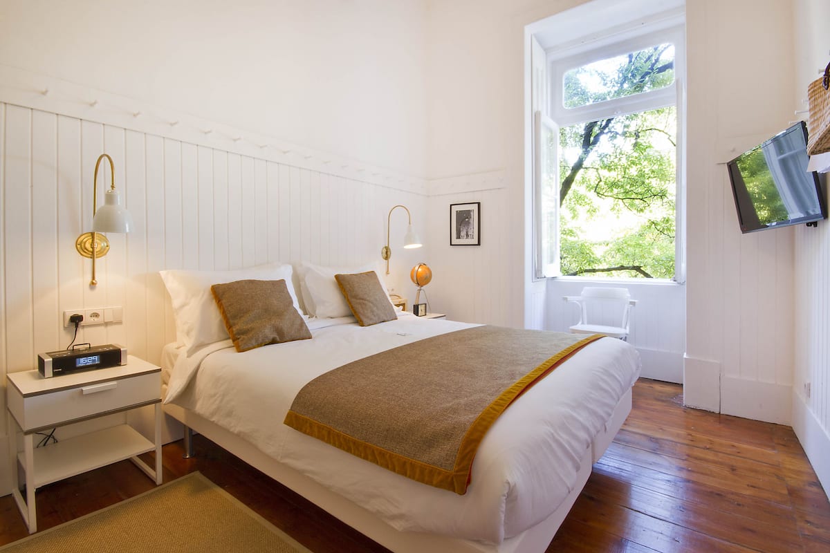 A neatly made bed with a white headboard, a bedside table with a lamp, and a window with a view of trees.