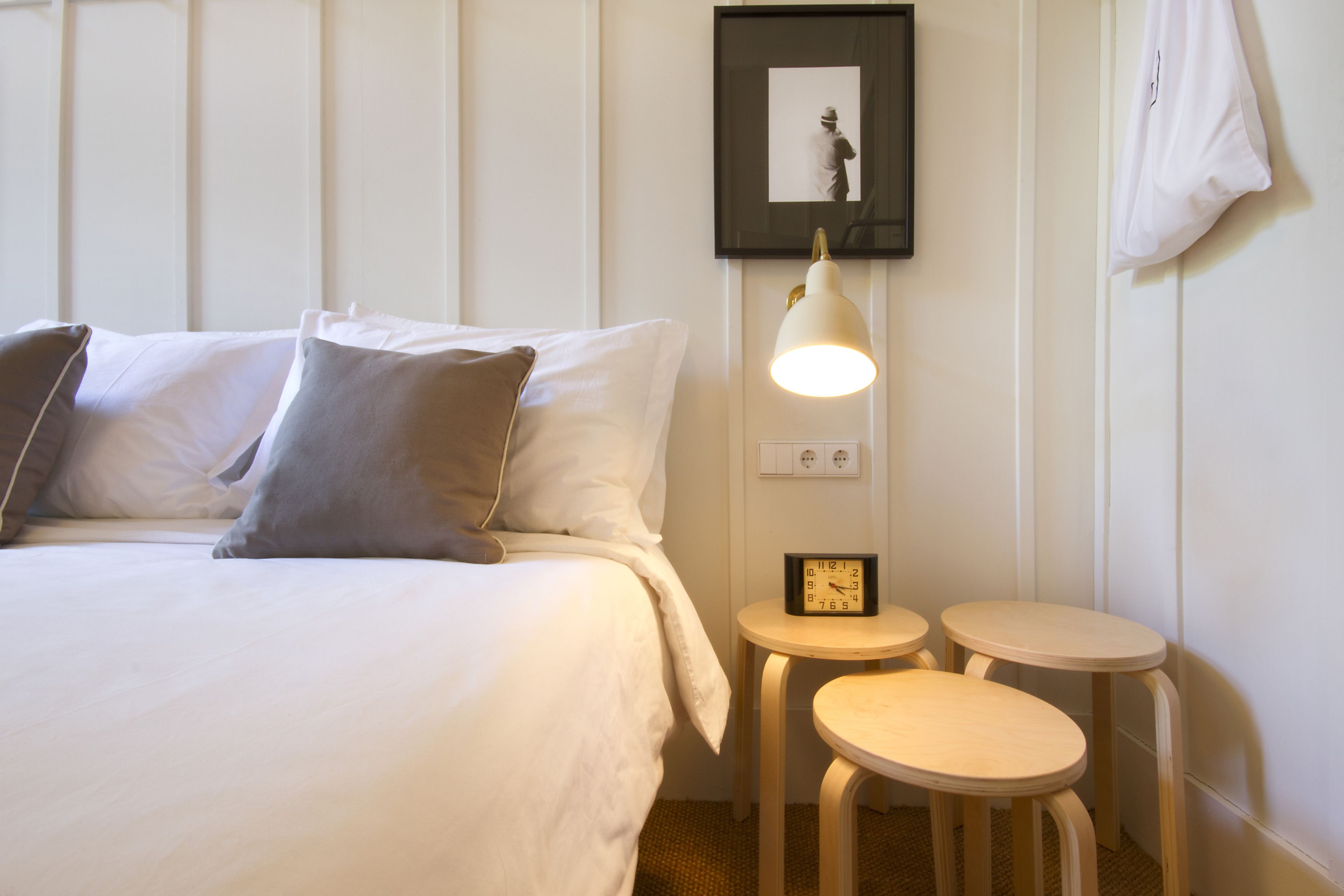 A neatly made bed with pillows, a bedside table with a clock, and two small stools.