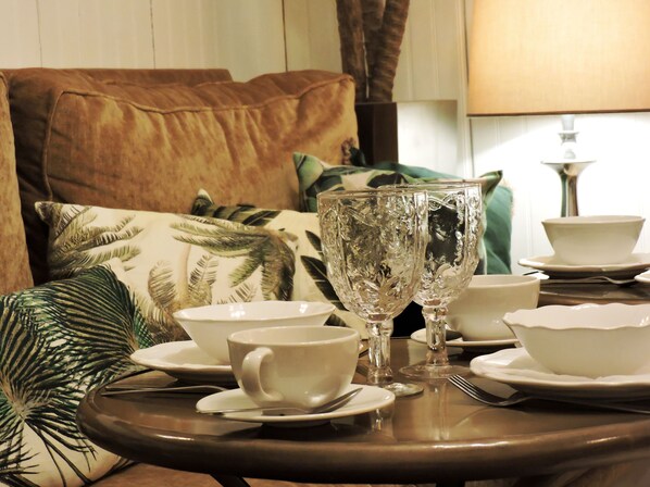 Una mesa de comedor dispuesta con vajilla blanca, dos copas de cristal y un cojín verde al fondo.