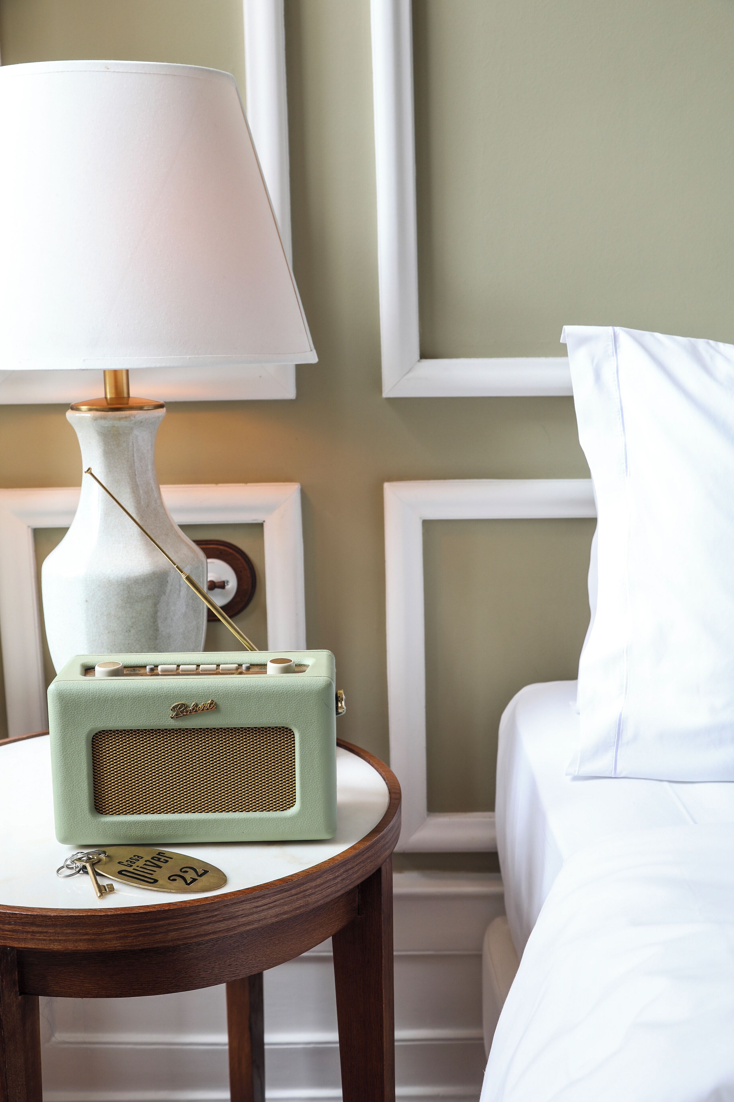 A vintage radio on a bedside table, a white lamp, and a white bed in a room with a green wall and framed artwork.