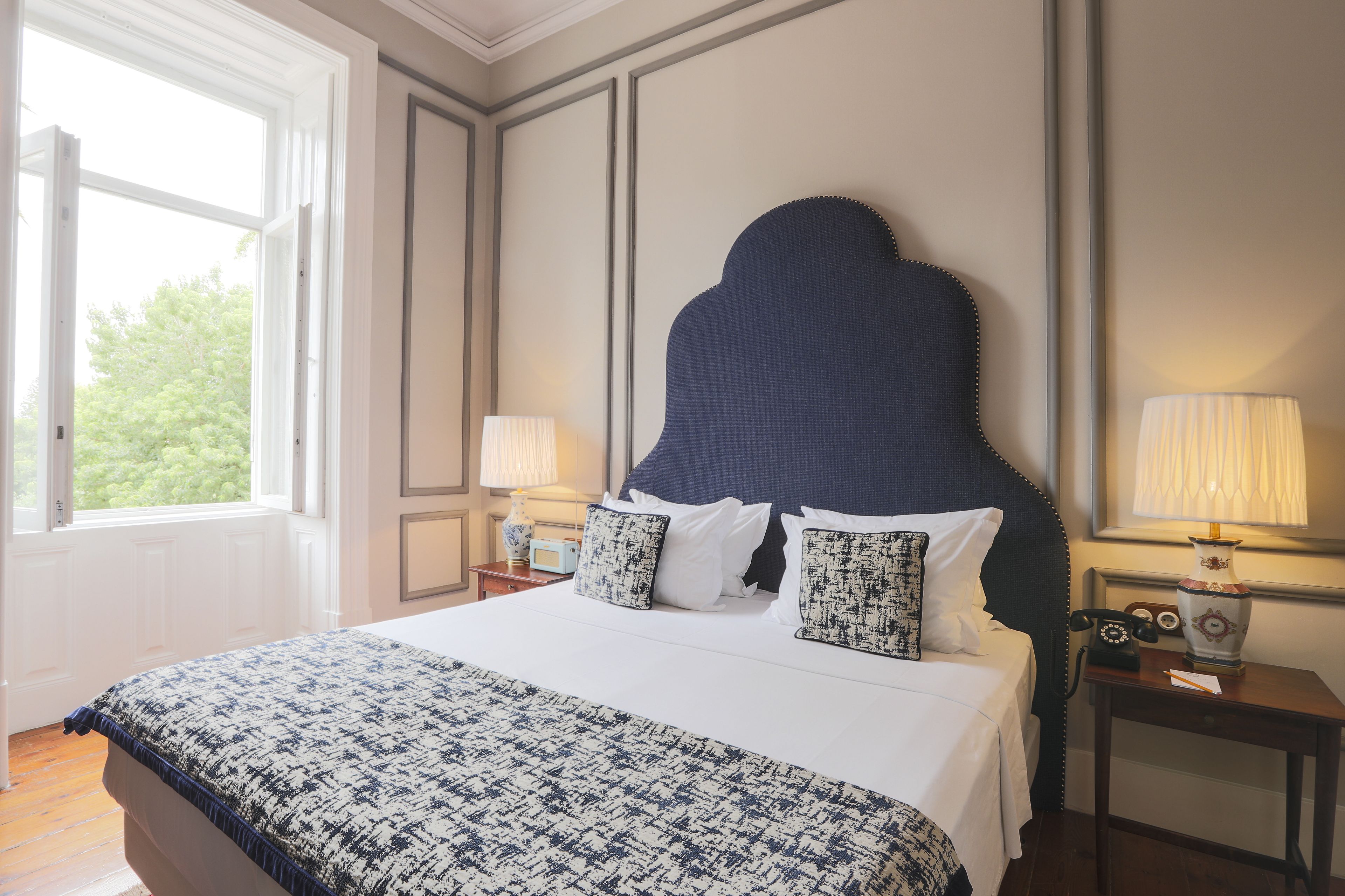 A neatly made bed with a patterned bedspread, a dark upholstered headboard, two bedside lamps, and a nightstand with a radio and a vase.