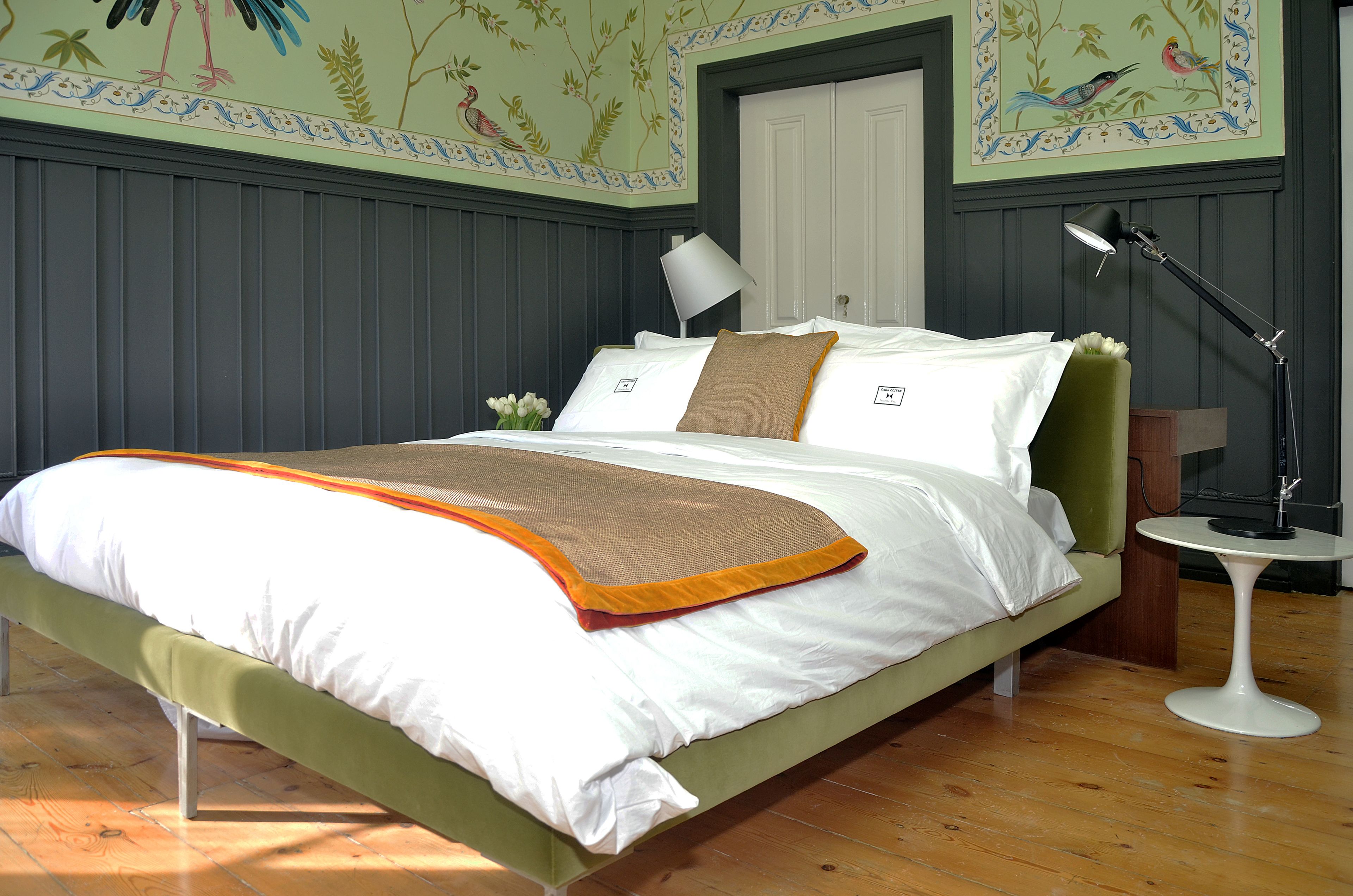 A neatly made bed with white and orange bedding, a green upholstered headboard, and a bedside table with a lamp.