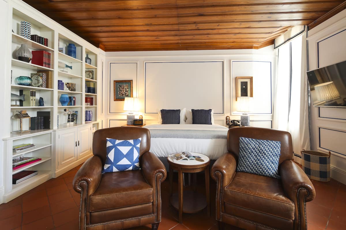 A cozy room with a bed, two leather armchairs, a small round table, and a bookshelf filled with books and decorative items.