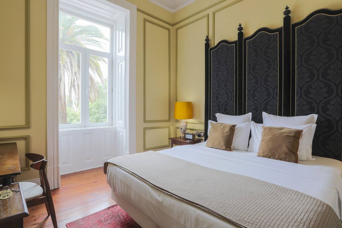 A hotel room with a large bed, a nightstand with a lamp, a chair, and a window with a view of palm trees.