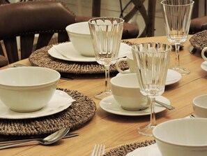 Una mesa de comedor dispuesta con cuencos blancos, platos y cristalería transparente, rodeada de individuales tejidos y sillas de cuero marrón.
