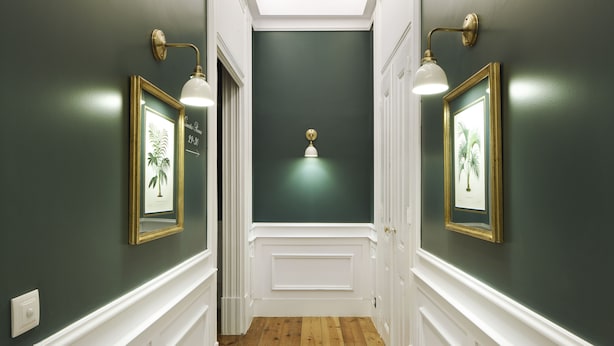 A hallway with dark green walls, white trim, and wooden flooring. There are framed pictures on the walls and a light fixture in the center.