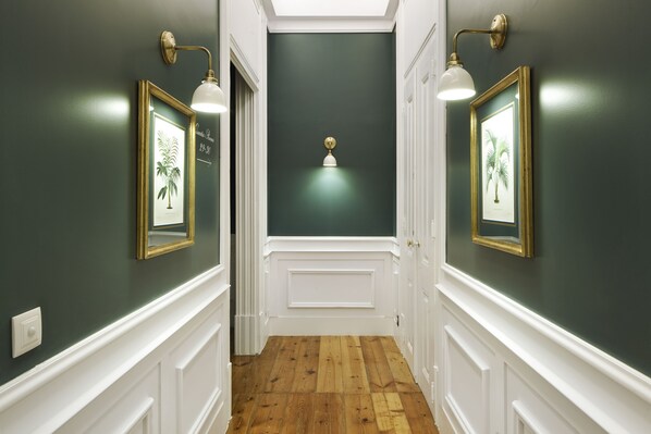 A hallway with dark green walls, white trim, and wooden flooring. There are framed pictures on the walls and a light fixture in the center.