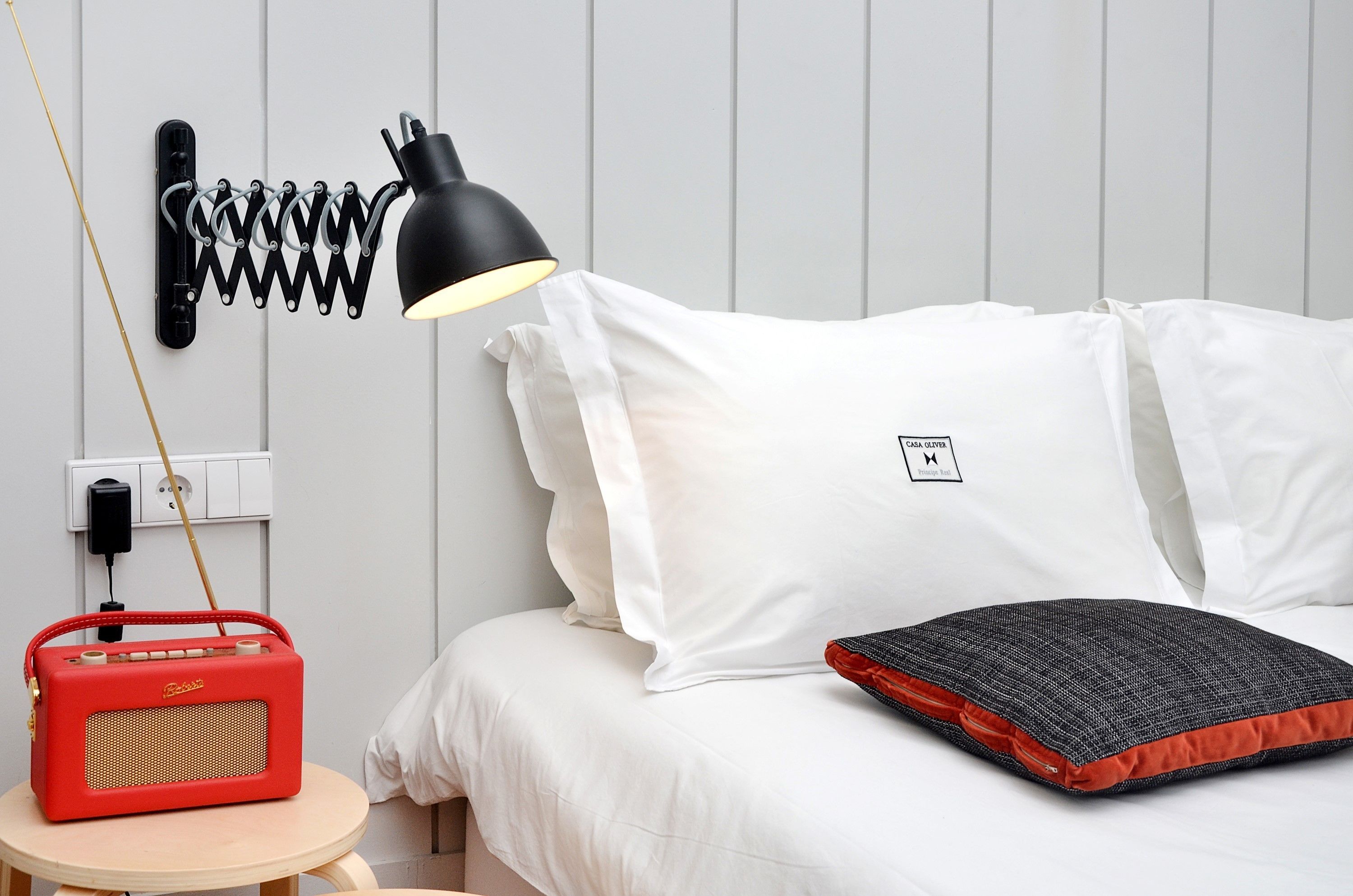 A neatly made bed with white linens, a black lamp, a red radio, and a black and red pillow.