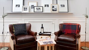 A room with two leather armchairs, a small table with a newspaper, a bottle, and two glasses. Above the chairs, there are framed black and white photographs on shelves.