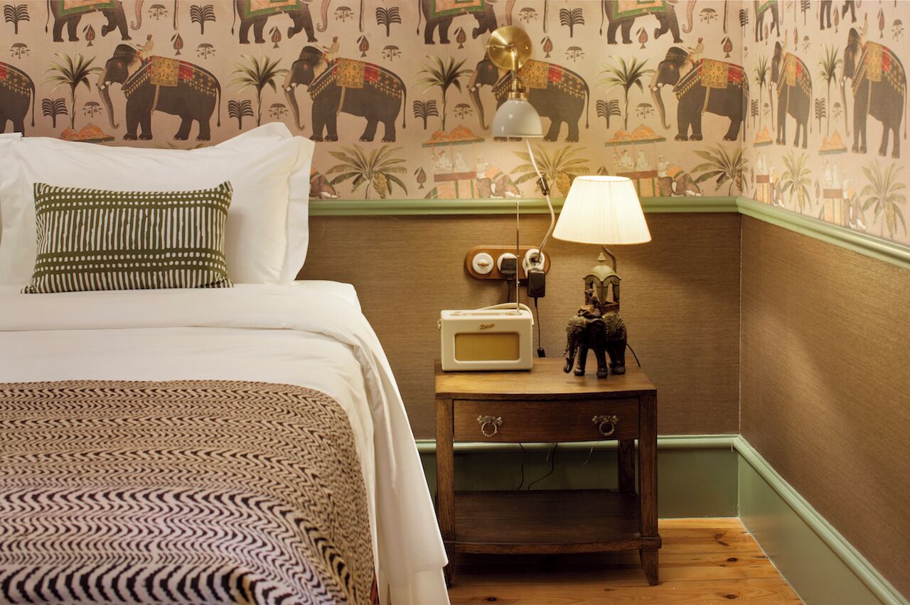 A bedroom with a bed, pillows, a nightstand with a lamp, and a wall with elephant patterns.
