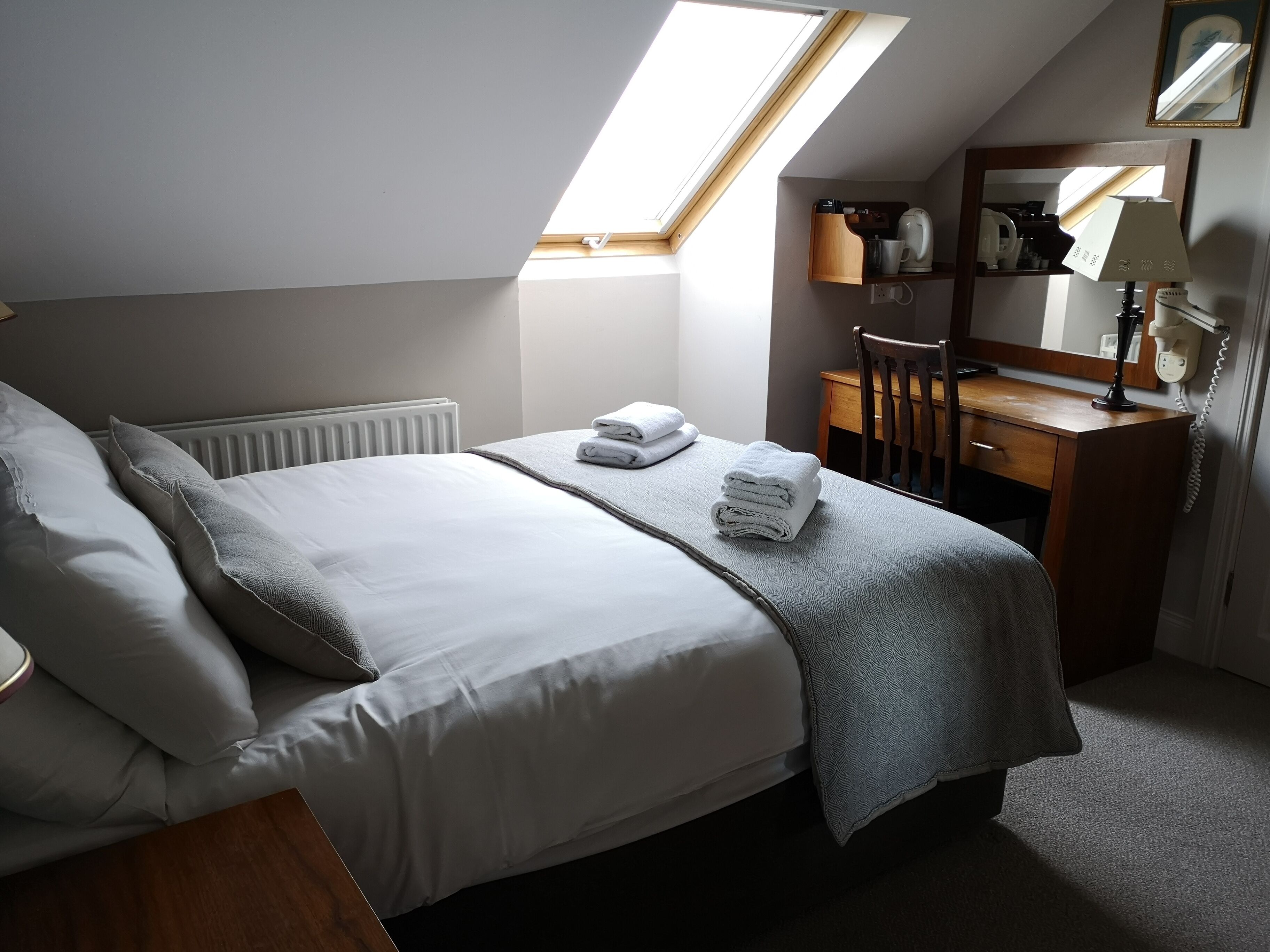 A bedroom with a skylight, a bed with white linens, a wooden dresser, and a mirror.