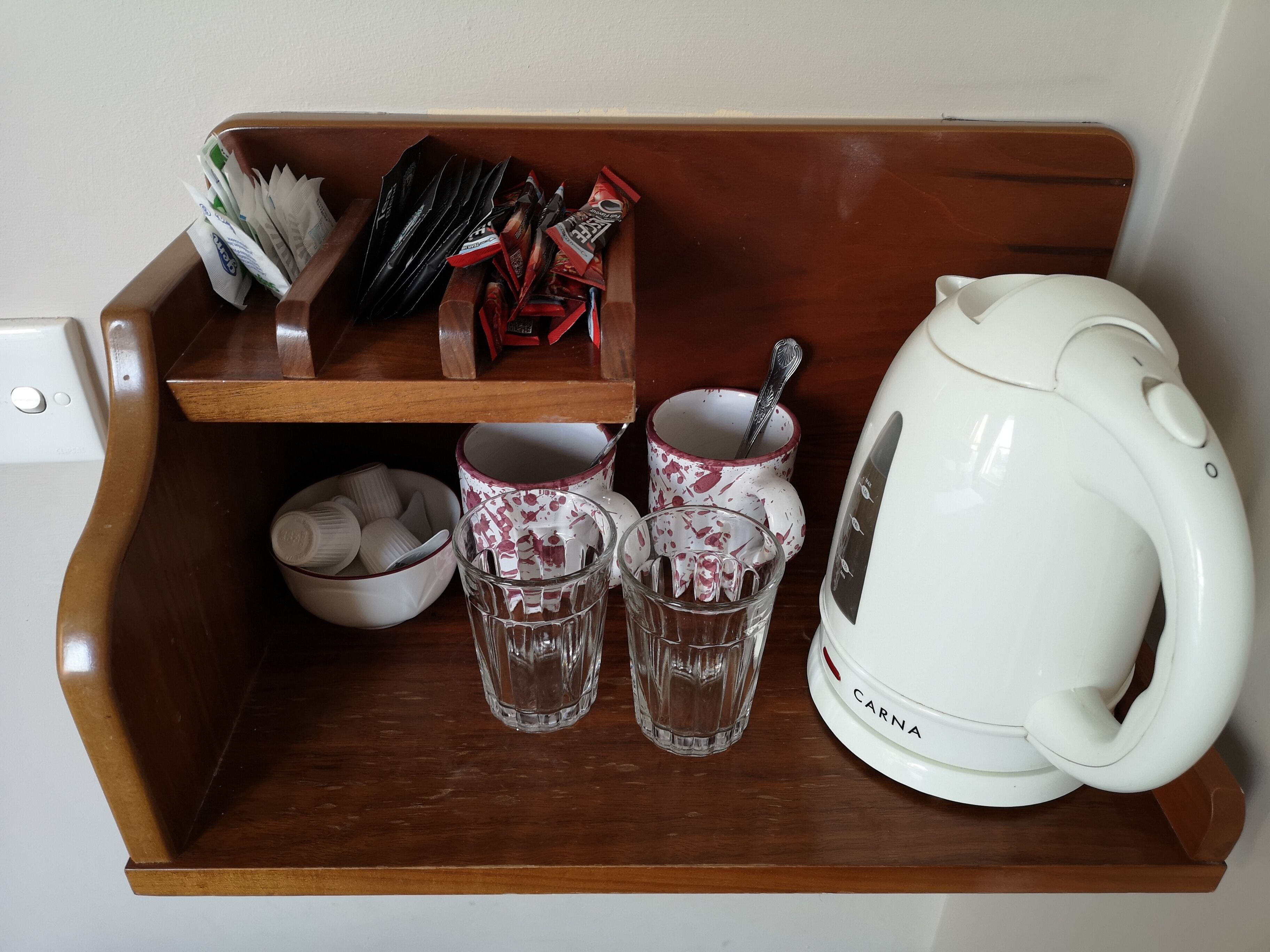 Una estantería de madera con un hervidor eléctrico blanco, dos vasos, un bol y una bandeja con varios objetos, incluyendo sobres y una cuchara.