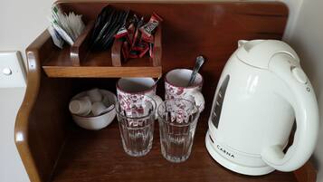 A wooden shelf with a white electric kettle, two glasses, a bowl, and a tray with various items including packets and a spoon.