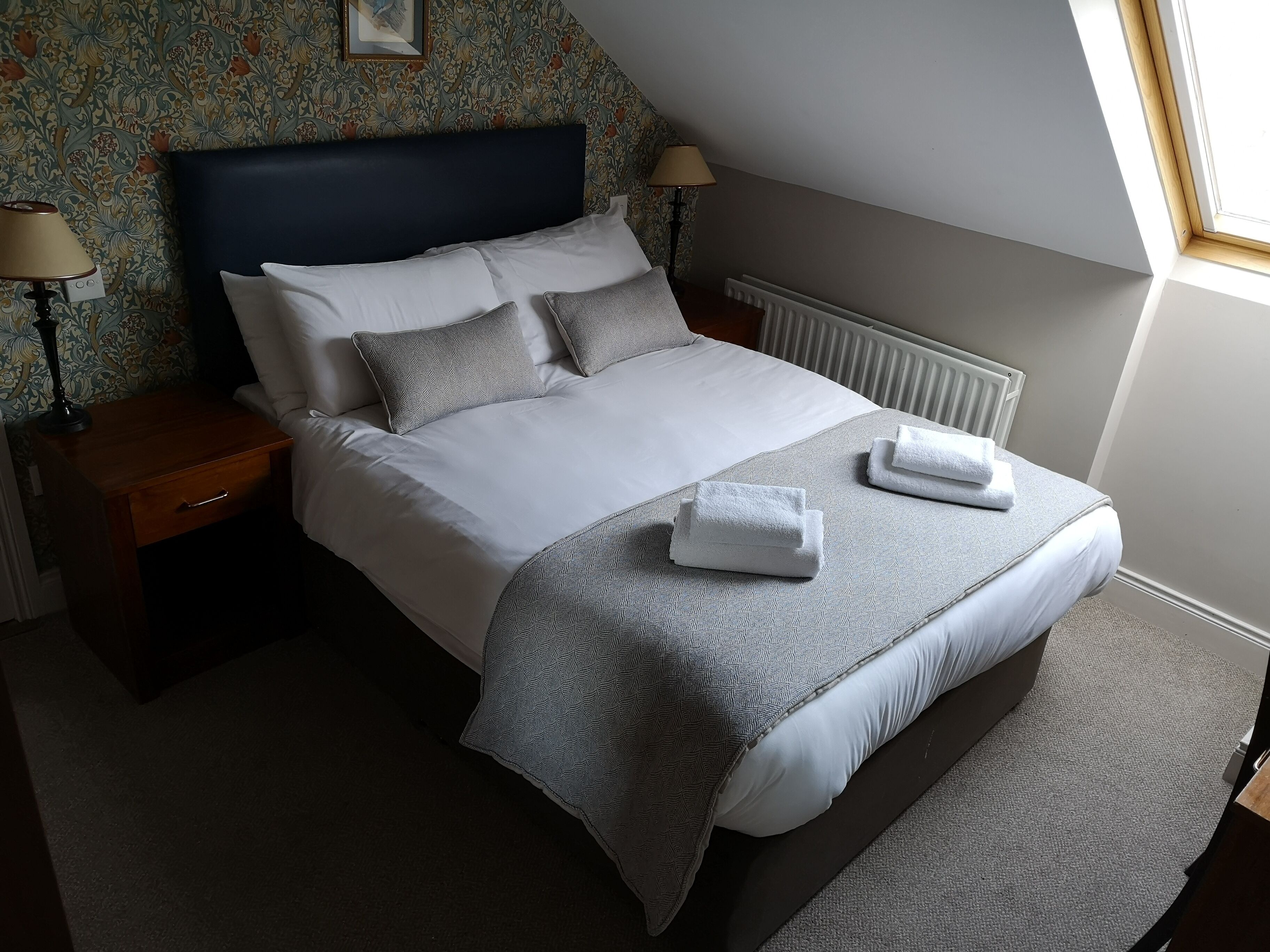 A neatly made bed with white linens and grey pillows, a bedside table with a lamp, and a radiator under the window.