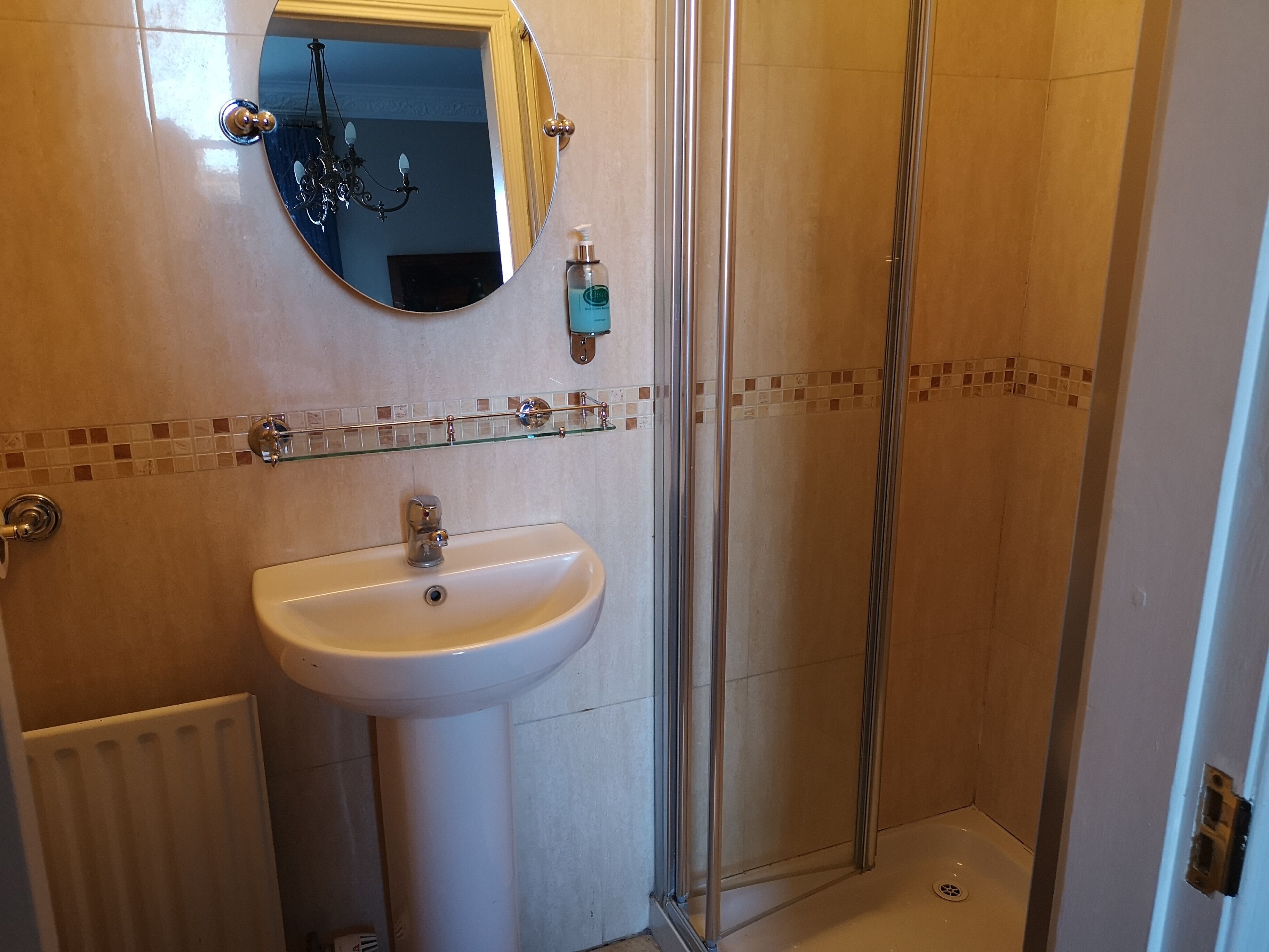 Un baño con un lavamanos blanco, un espejo redondo, una cabina de ducha con mampara de vidrio y un salpicadero de azulejos.