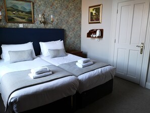 Una cama doble con sábanas blancas y almohadones grises, una mesita de noche con un hervidor blanco, y una puerta con pomo de latón.