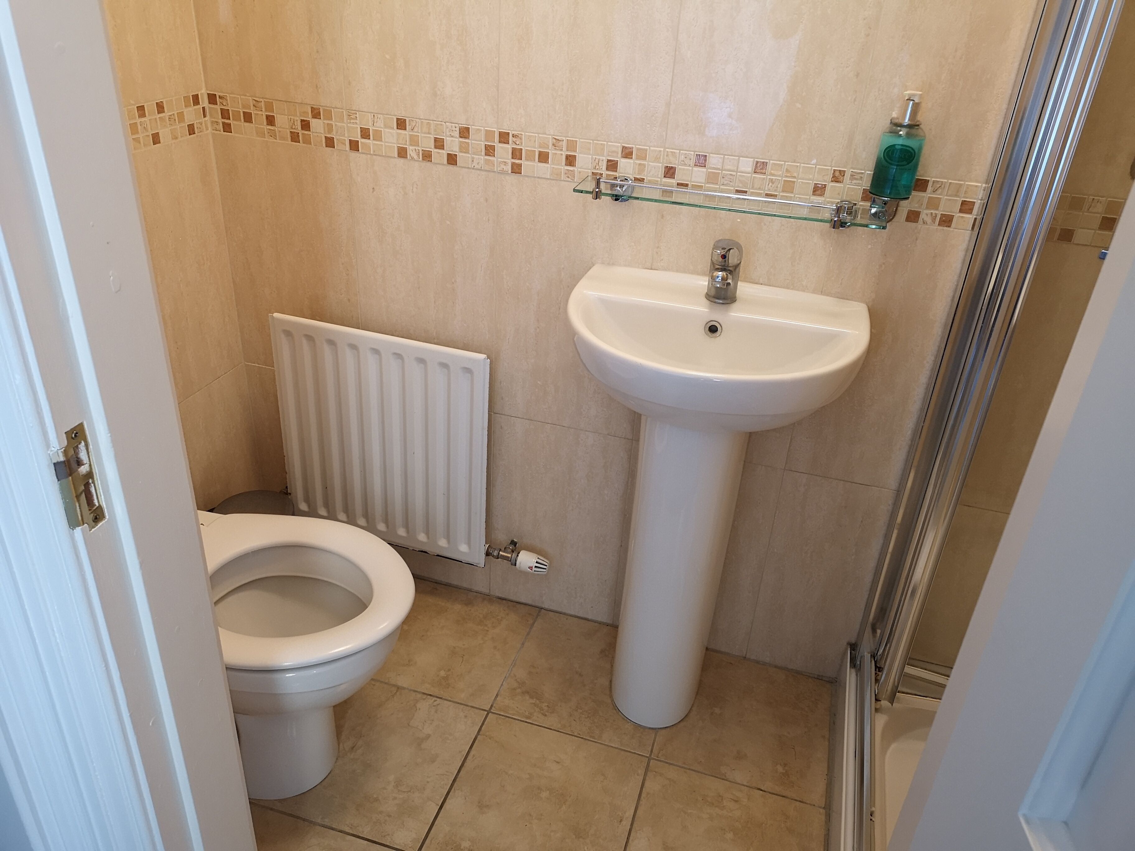 Un baño con inodoro, lavamanos pedestal, repisa de vidrio con artículos de aseo y radiador.