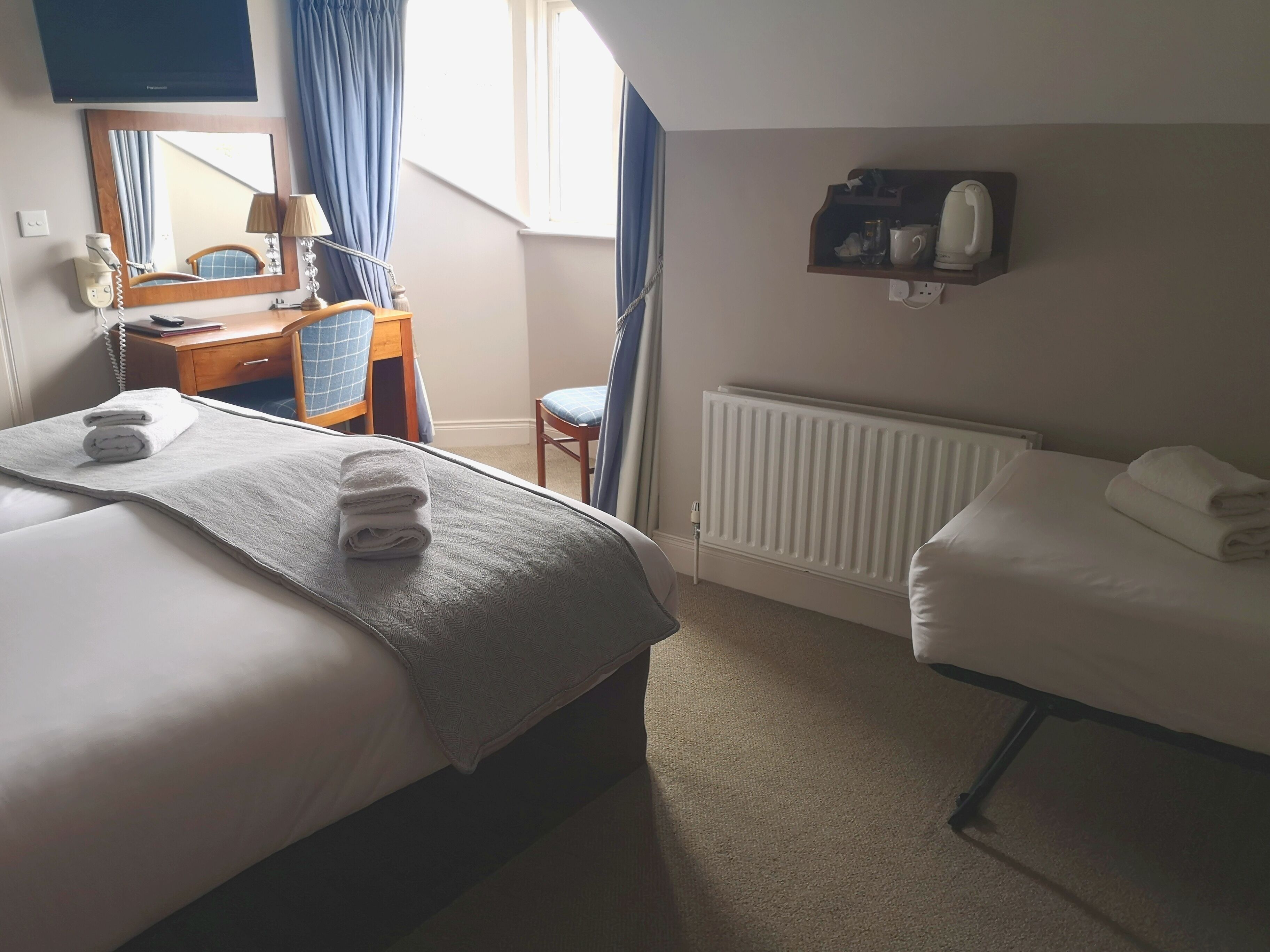 Habitación de hotel con dos camas, un escritorio, una silla, un espejo, un radiador y una repisa con una tetera y una taza.