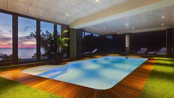 Una piscina interior moderna con terraza de madera y césped artificial, con amplios ventanales que ofrecen vistas del atardecer.