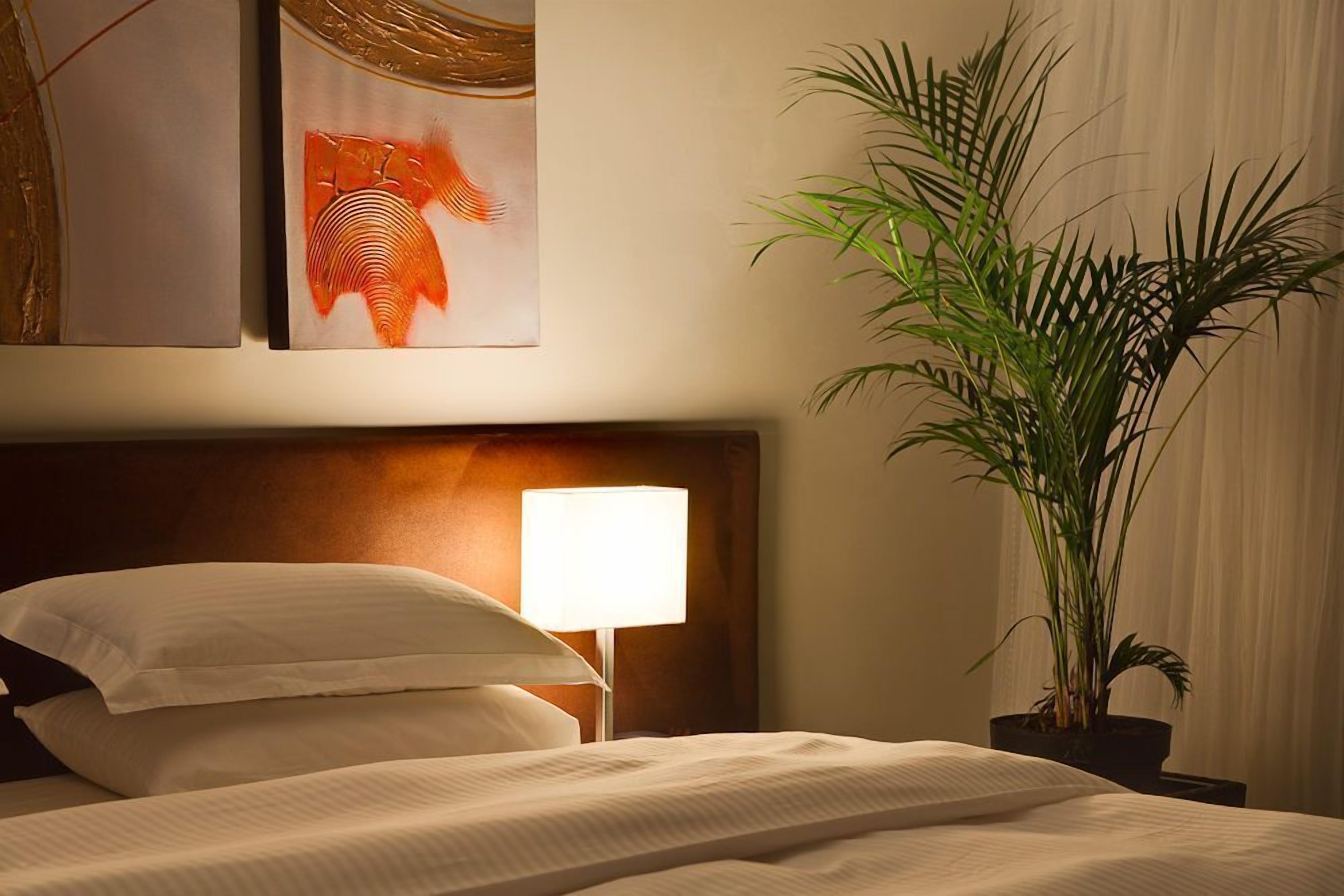 A neatly made bed with white linens, a bedside lamp, and a large potted plant.
