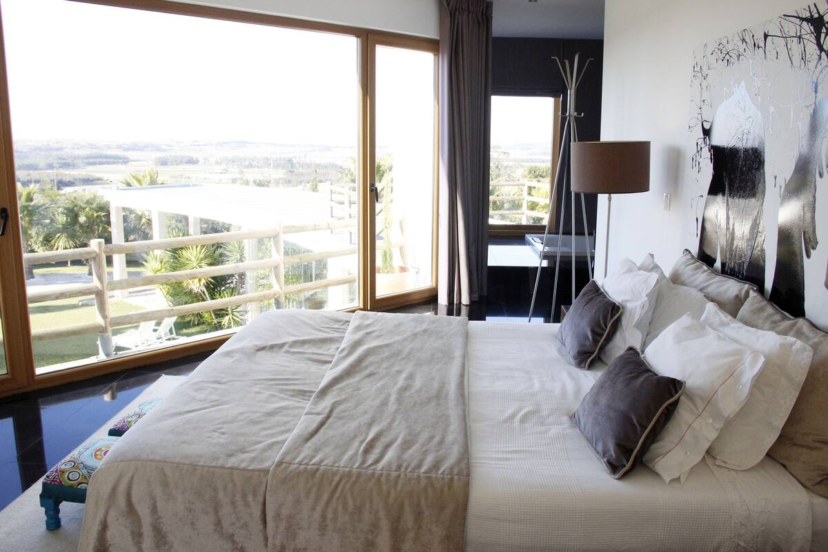 A large bed with multiple pillows, a view of the outdoors through a sliding door, and a painting on the wall.