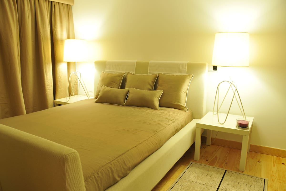 A neatly made bed with a beige bedspread and pillows, two bedside lamps, a small white nightstand with a pink object, and a wooden floor.