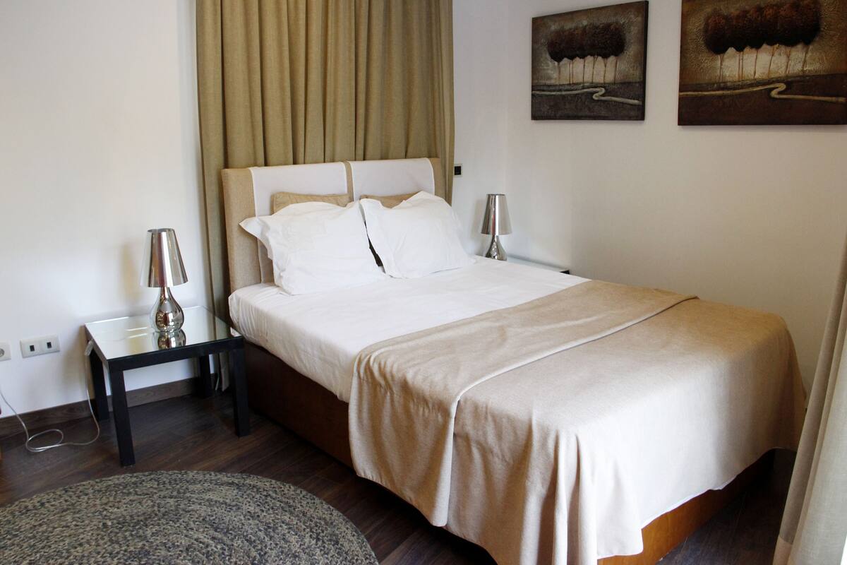 A neatly made bed with a beige bedspread and pillows, a bedside table with a lamp, and a wall with two abstract paintings.